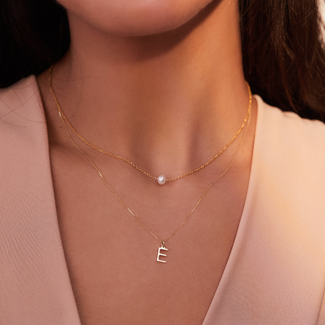 A brunette woman wearing a gold single pearl choker and solid gold individual initial charm 'E' around her neck