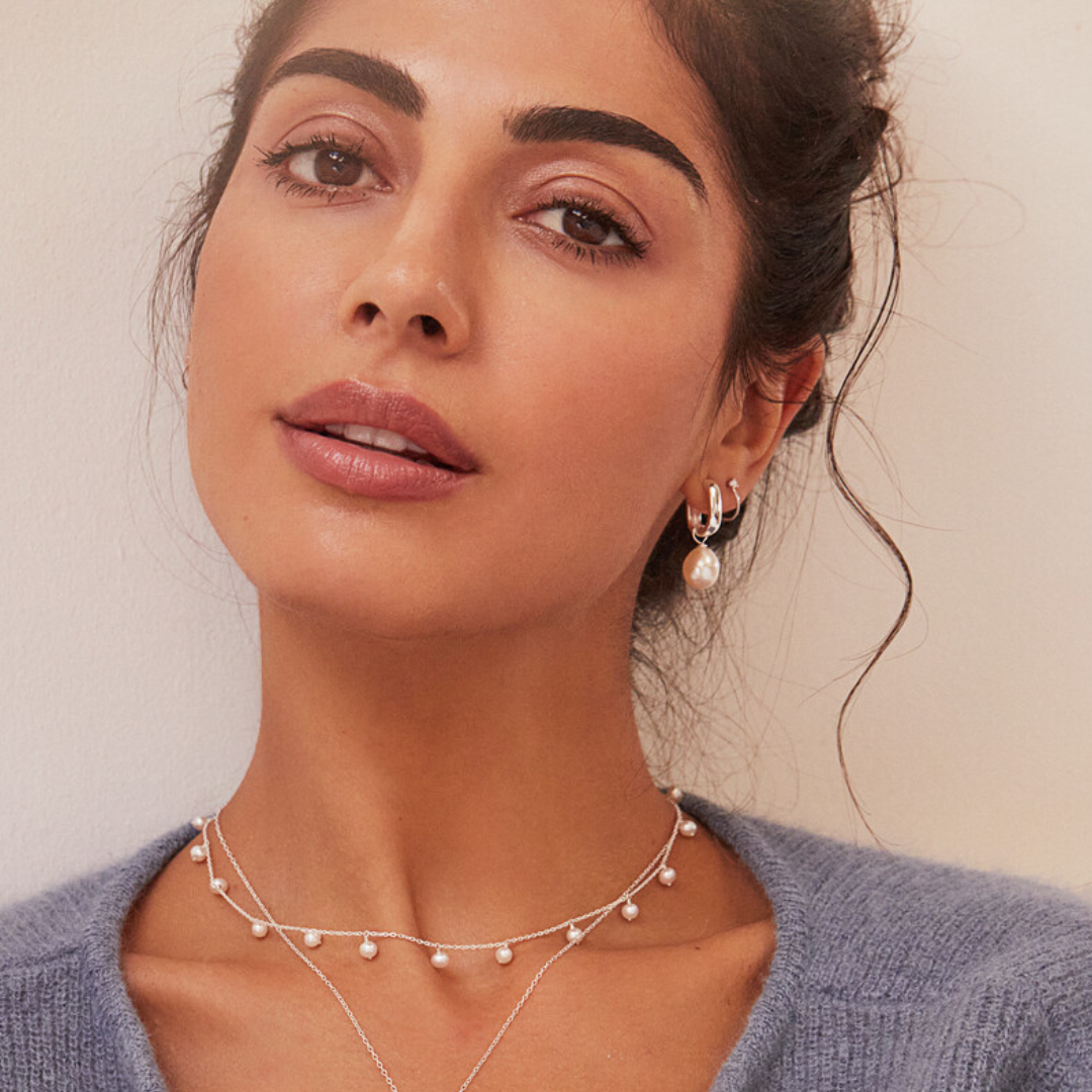 A woman wearing a silver diamond style lobe hoop stud earring and a silver thick squared hoop pearl drop earring in her ear lobe with a silver pearl choker around her neck  