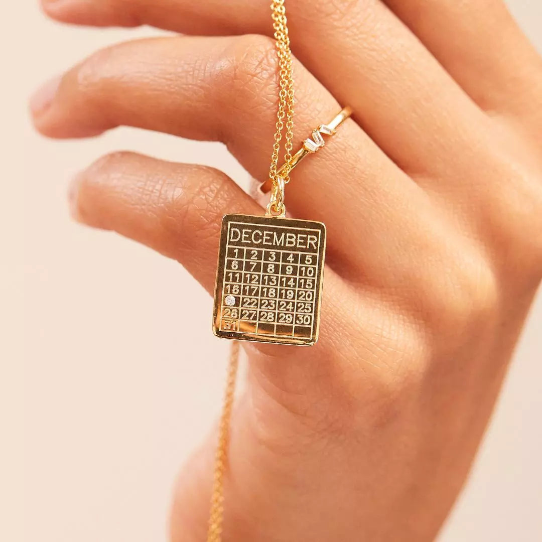 Gold special date calendar necklace held up by a hand with a gold diamond style baguette eternity ring on one finger
