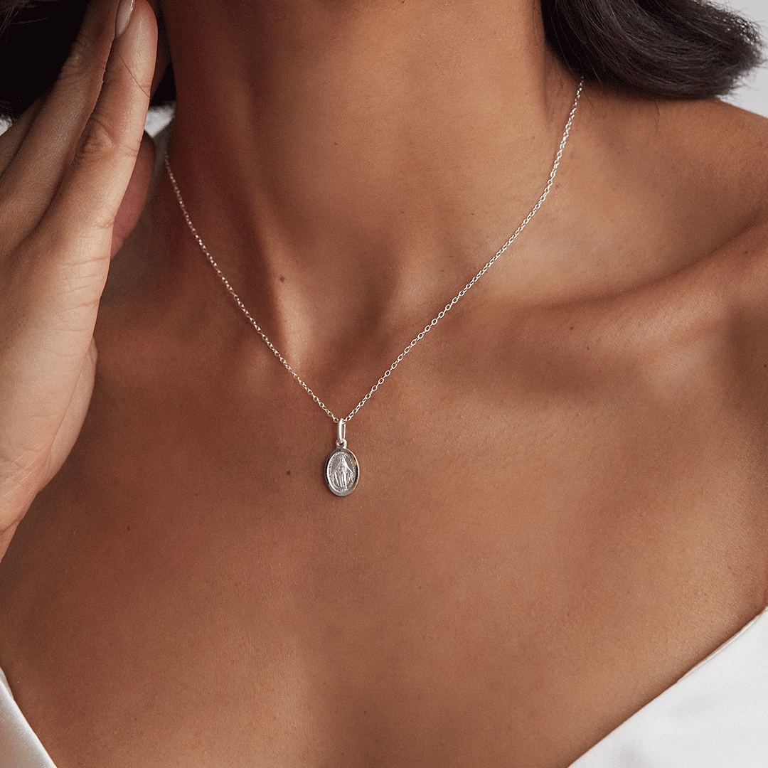 Silver small virgin mary necklace around the neck of a woman wearing a white strappy top