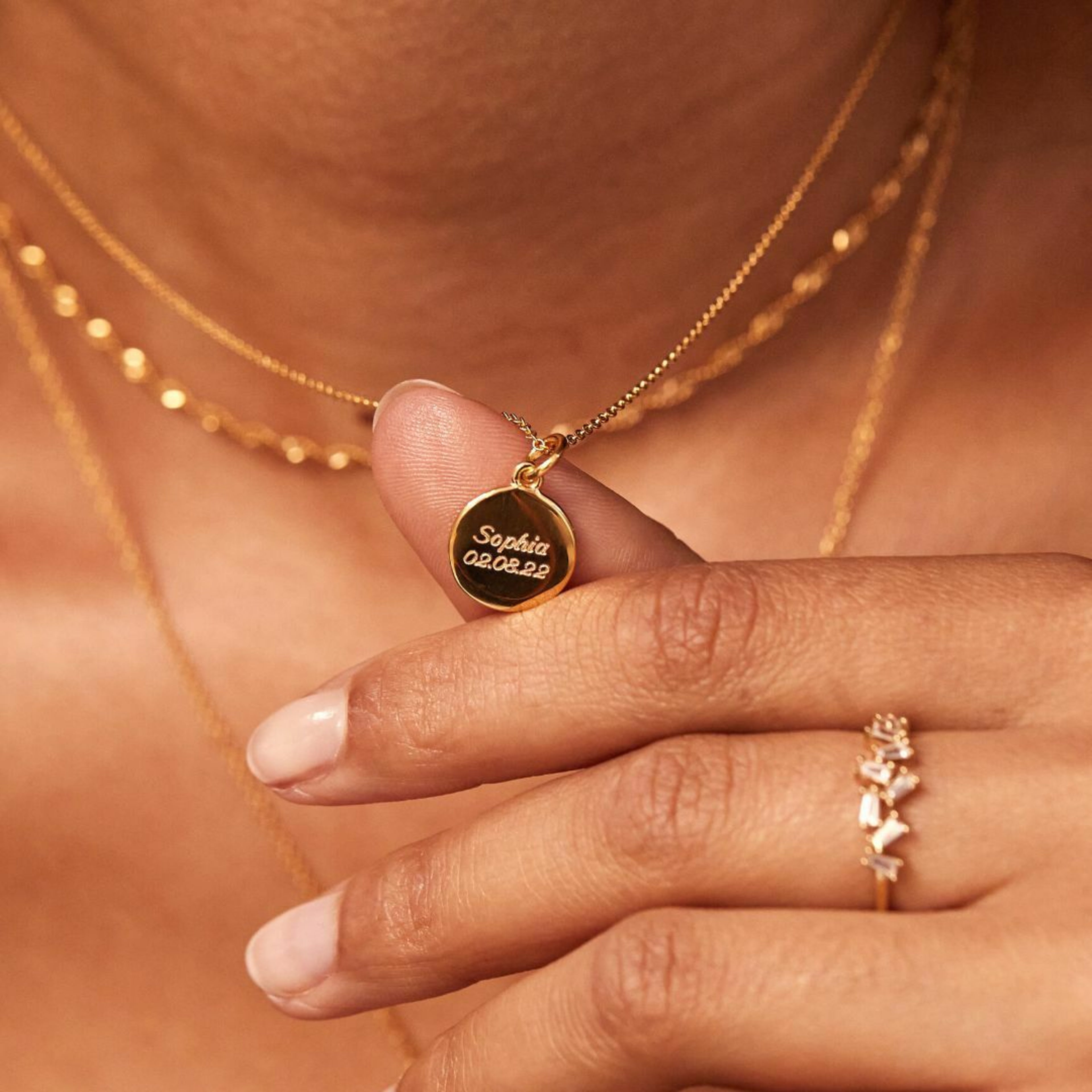 Gold small round engraved disc necklace with the words 'Sophia 02.08.22' engraved around woman's neck held up by her thumb