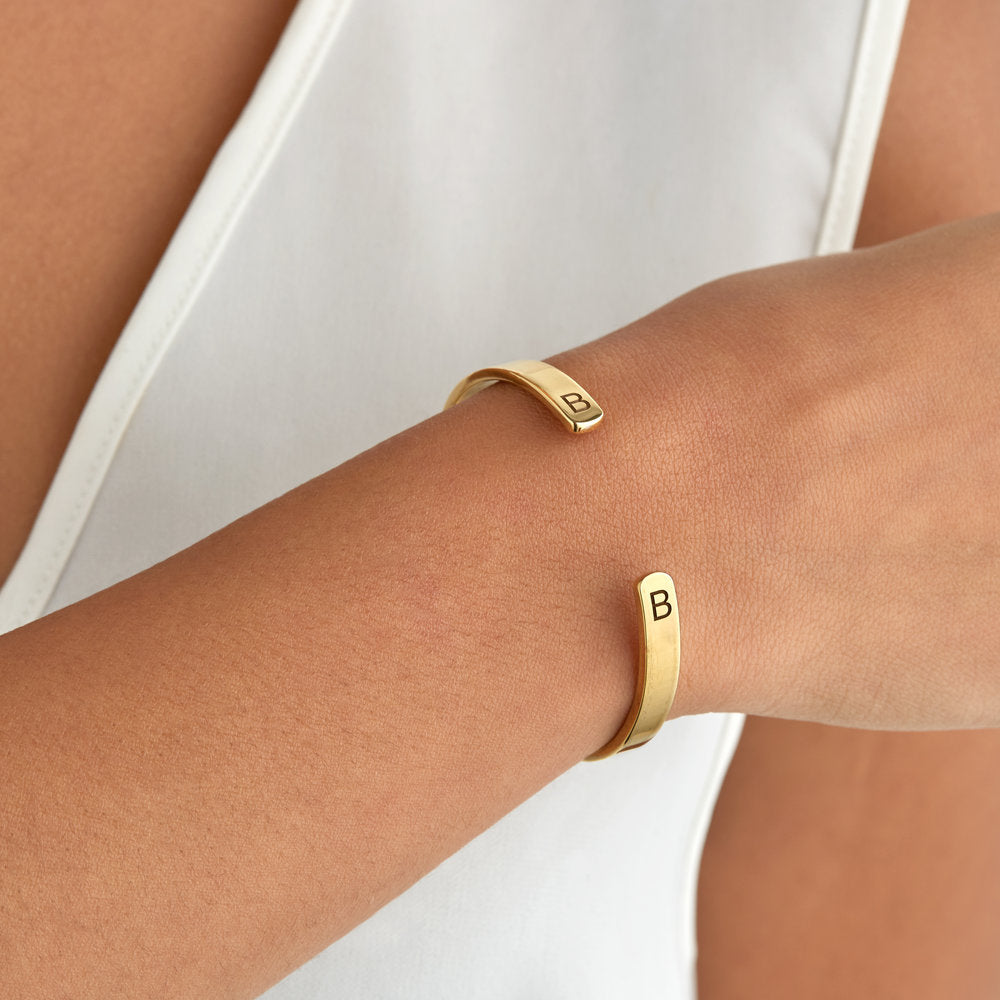 Gold thick engraved bangle with the letter 'B' around a wrist with a white top in the background