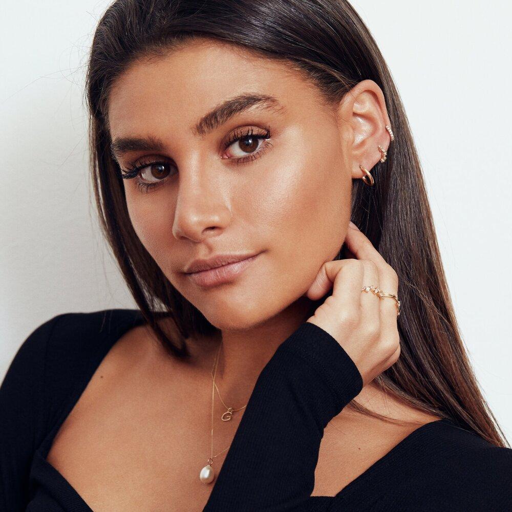 A brunette woman wearing a gold large single pearl necklace around her neck and a gold diamond style baguette ring on her finger and gold diamond style huggie hoop earrings in her ear lobe, looking at the camera with her hair tucked behind her ear