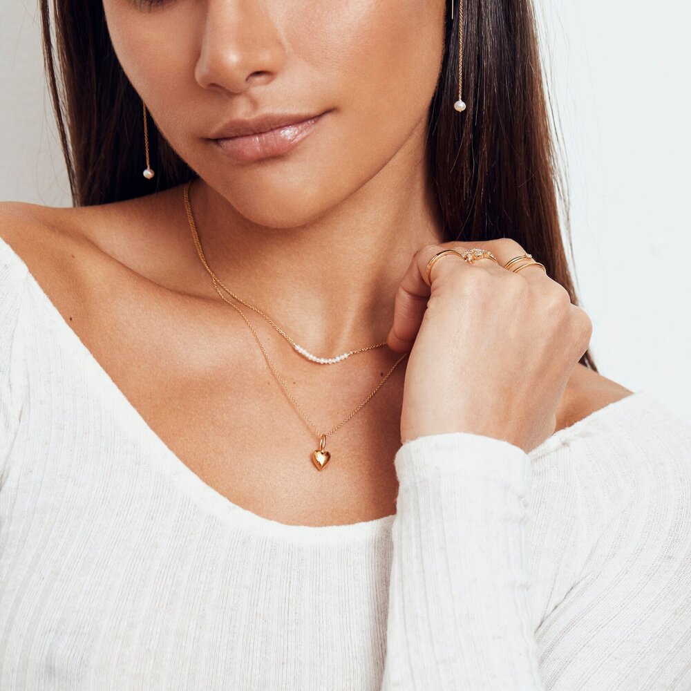 A brunette woman wearing a gold pearl cluster choker layered with a gold heart pendant necklace around her neck and gold pearl drop ear threaders in her ear lobes 