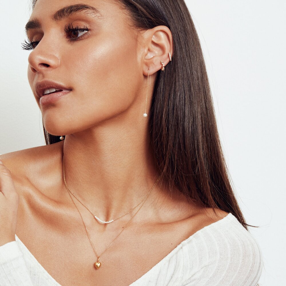 Gold pearl drop ear threaders in the ears of a brunette woman looking to the side also wearing gold diamond style jagged huggie hoop earrings in her ear lobe and a gold heart pendant necklace and gold pearl cluster choker around her neck