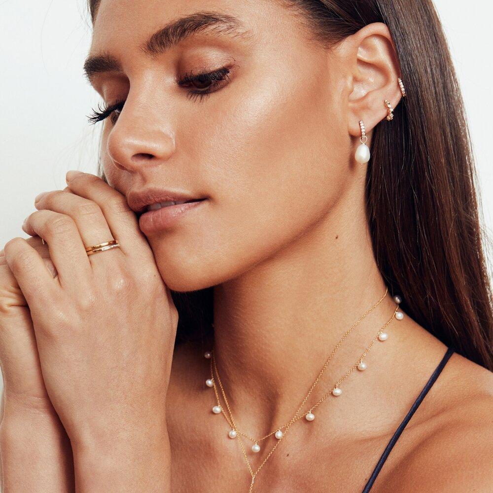 Woman looking down wearing a gold pearl drop choker layered with a gold necklace around her neck wearing a gold diamond style large pearl drop hoop earring and two gold diamond style huggie hoop earrings in her ear lobe