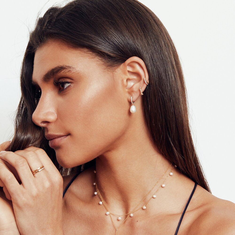 A woman wearing a gold diamond style huggie hoop earring and a gold diamond style large pearl drop hoop earring in her ear lobe and a gold pearl drop choker layered with a gold necklace around her neck  