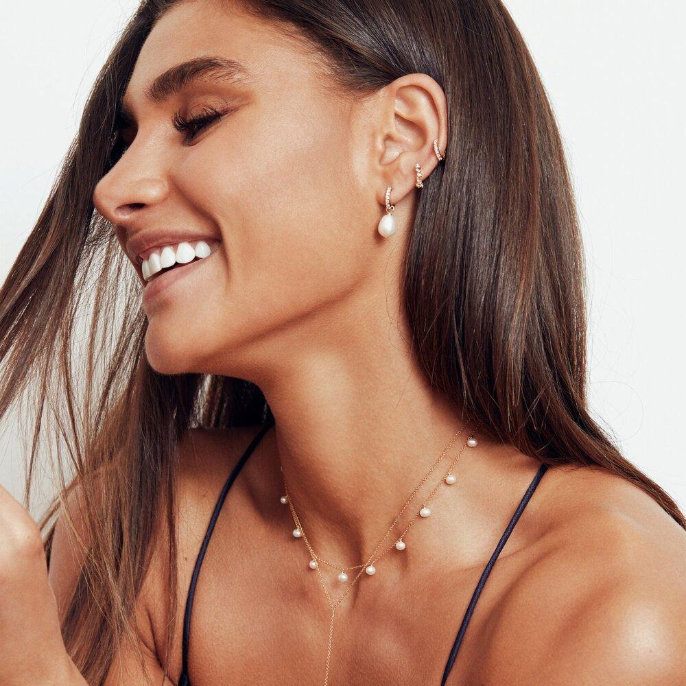 Gold pearl drop choker layered with a gold necklace around a neck of a woman wearing a gold diamond style large pearl drop hoop earring and a gold diamond style huggie hoop earring in her ear lobe