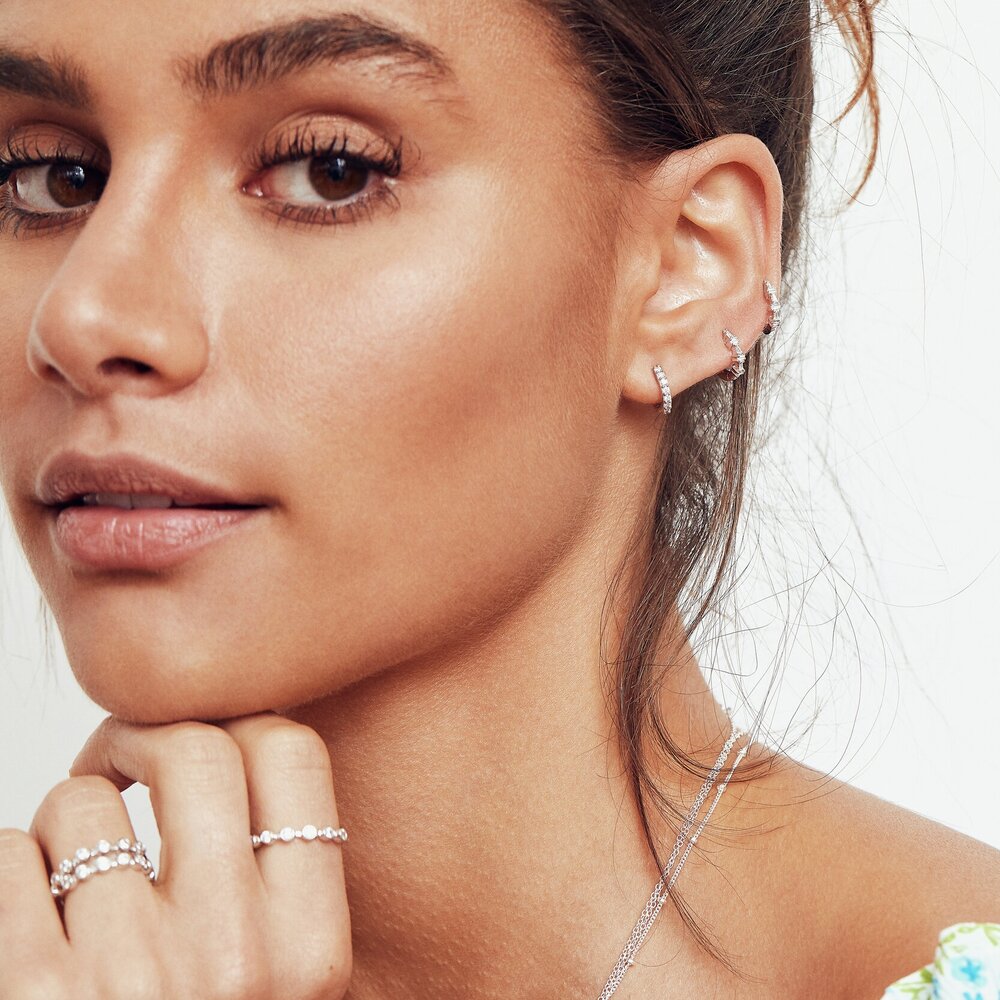 A woman wearing three silver diamond style round eternity rings on her fingers and three diamond style cluster huggie hoop earrings in her ear lobes 
