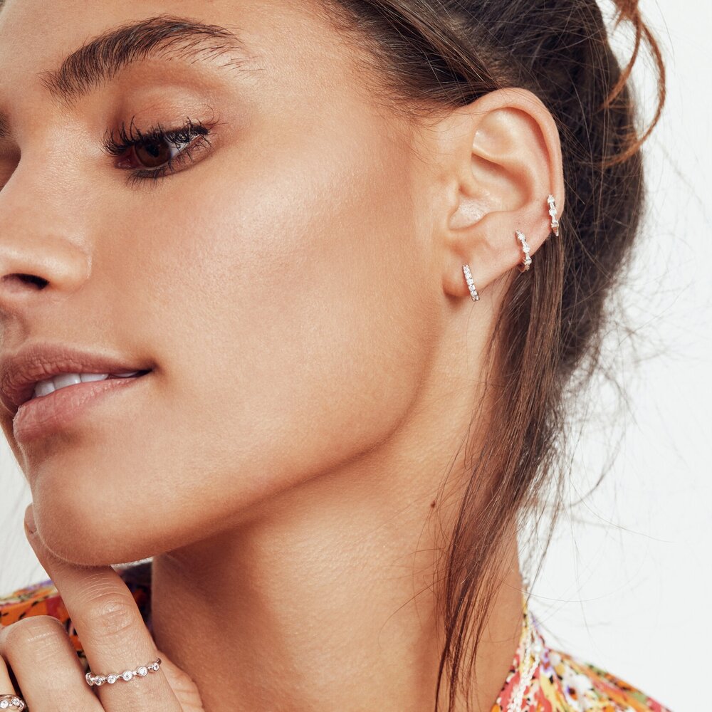 A close up of a woman with a silver diamond style round eternity ring on her finger and three diamond style cluster huggie hoop earrings in her ear