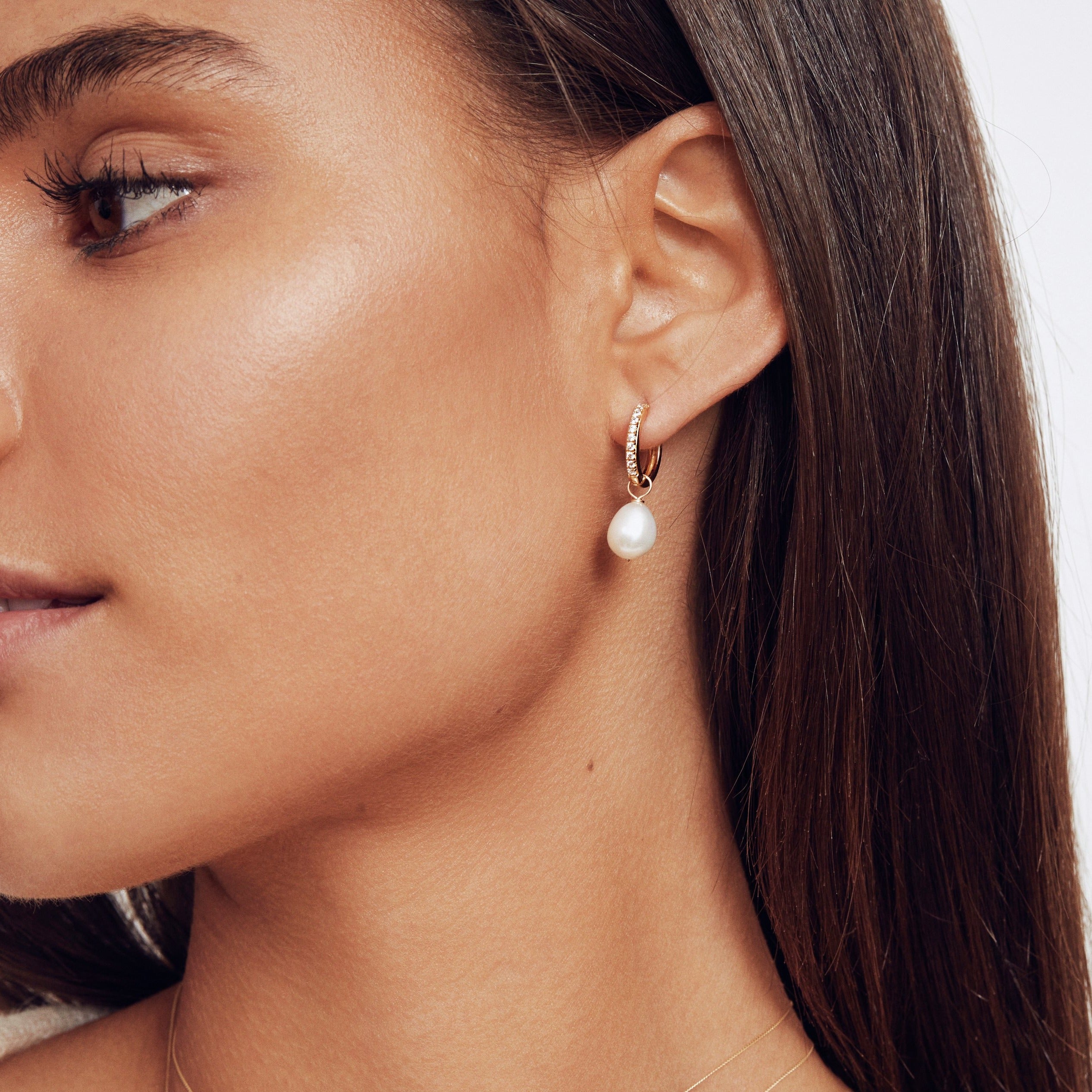 A silver diamond style large pearl drop hoop earring in the ear lobe of a brunette woman