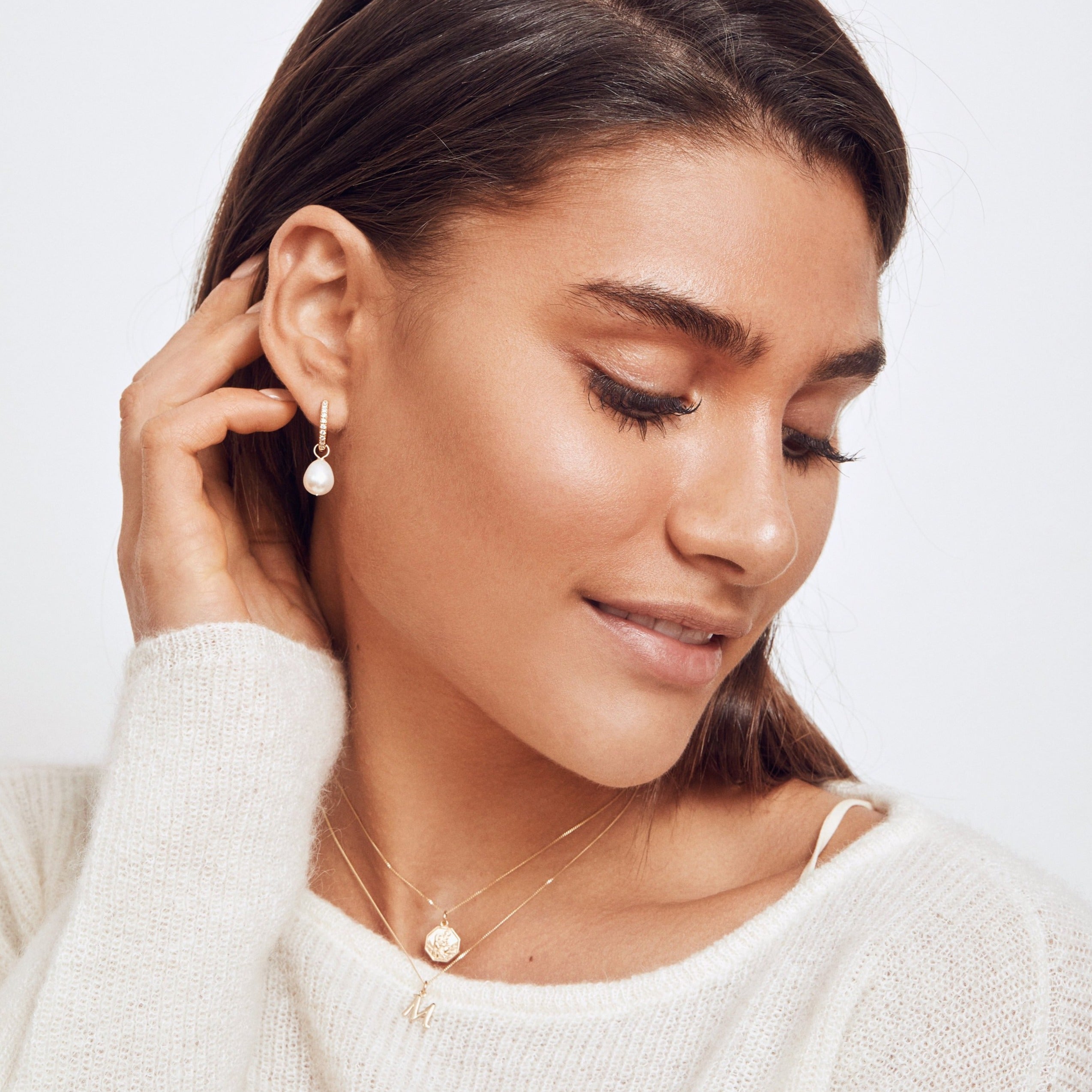 A gold diamond style large pearl drop hoop earring in the ear lobe of a woman wearing a gold individual curve initial charm 'M' around her neck
