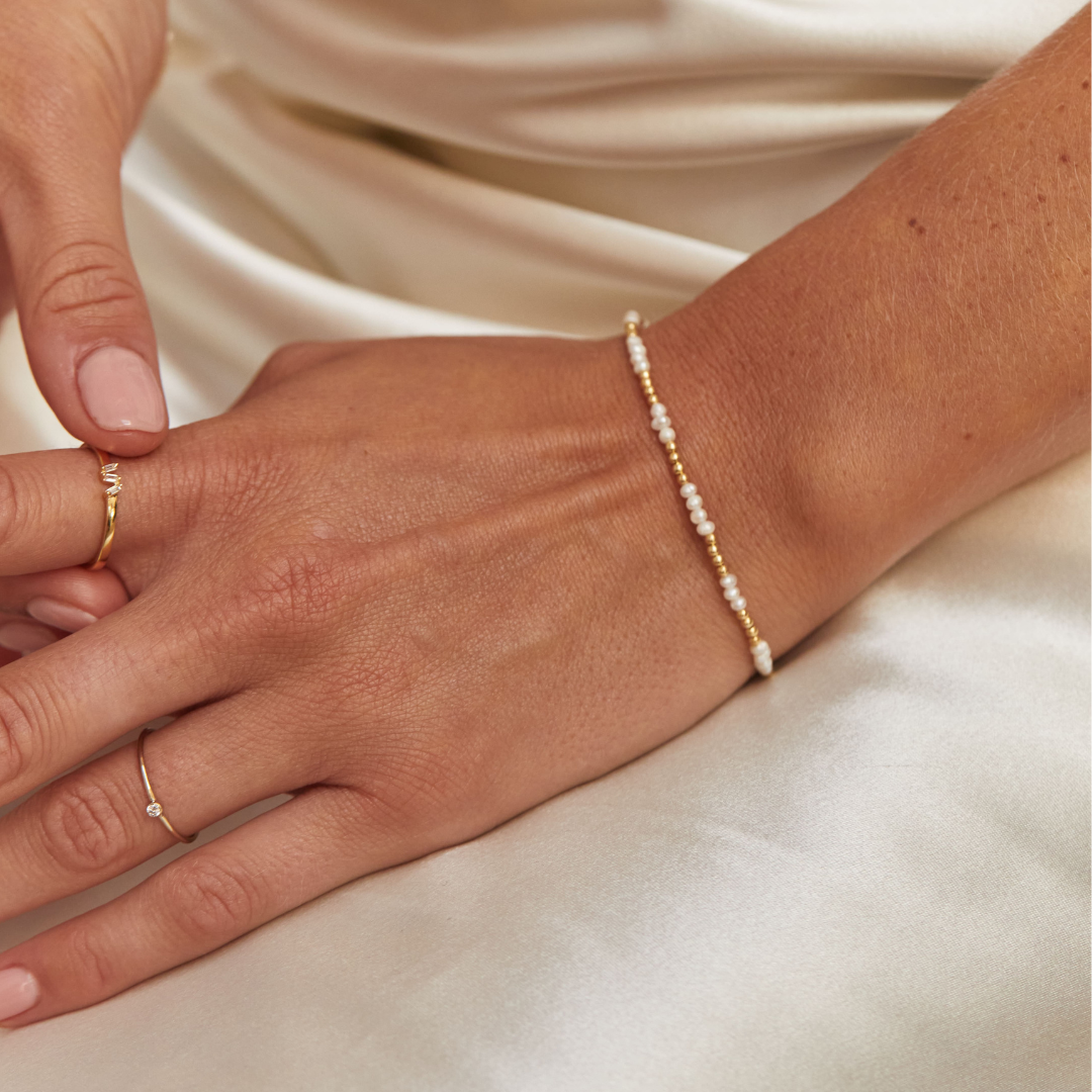 Gold beaded mini pearl bracelet around a wrist paired with a gold diamond style three baguette ring on one finger and a gold thin diamond style stacking ring on another finger