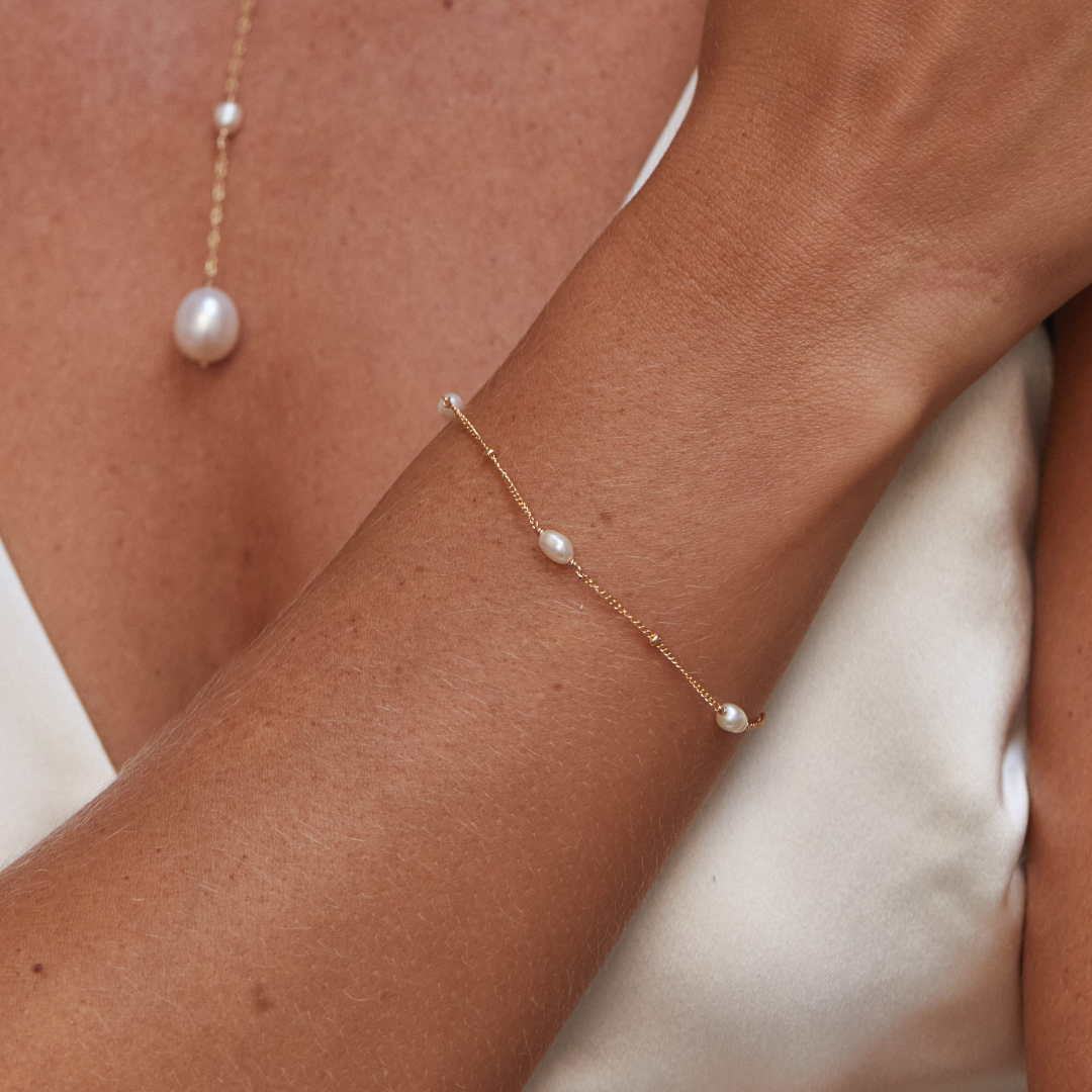 Close up gold seed pearl satellite bracelet on a wrist of a woman wearing white and a gold pearl lariat necklace