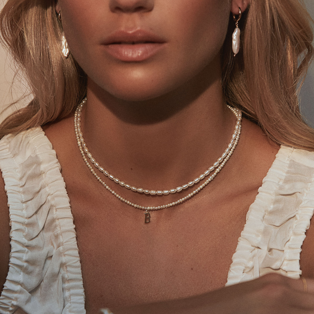 Close up of a blonde woman wearing a gold small pearl initial letter choker and gold seed pearl choker around her neck with gold baroque pearl shard hoop earrings in her ear lobes