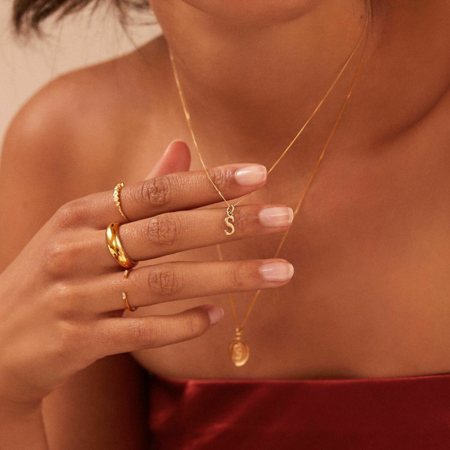 Solid gold curve initial letter necklace 'S' around the neck of a woman holding it up with the back of her fingers that are wearing a gold plain dome ring, a gold diamond style eternity ring and a gold diamond style baguette gap ring on her fingers