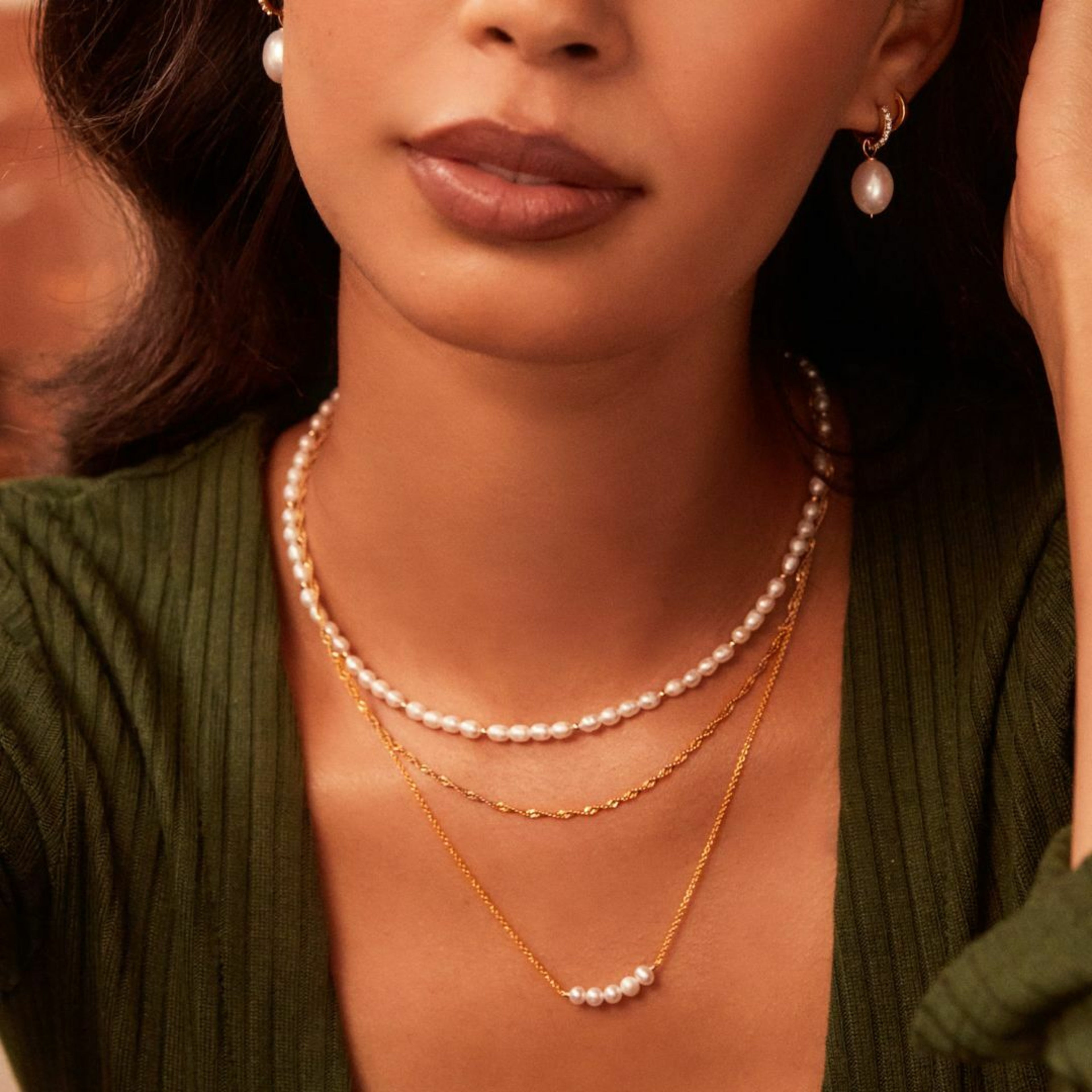 Close up gold beaded seed pearl choker on a neck layered with a gold pearl cluster necklace, a twisted gold chain necklace and gold diamond style large pearl drop hoop earrings in her ears
