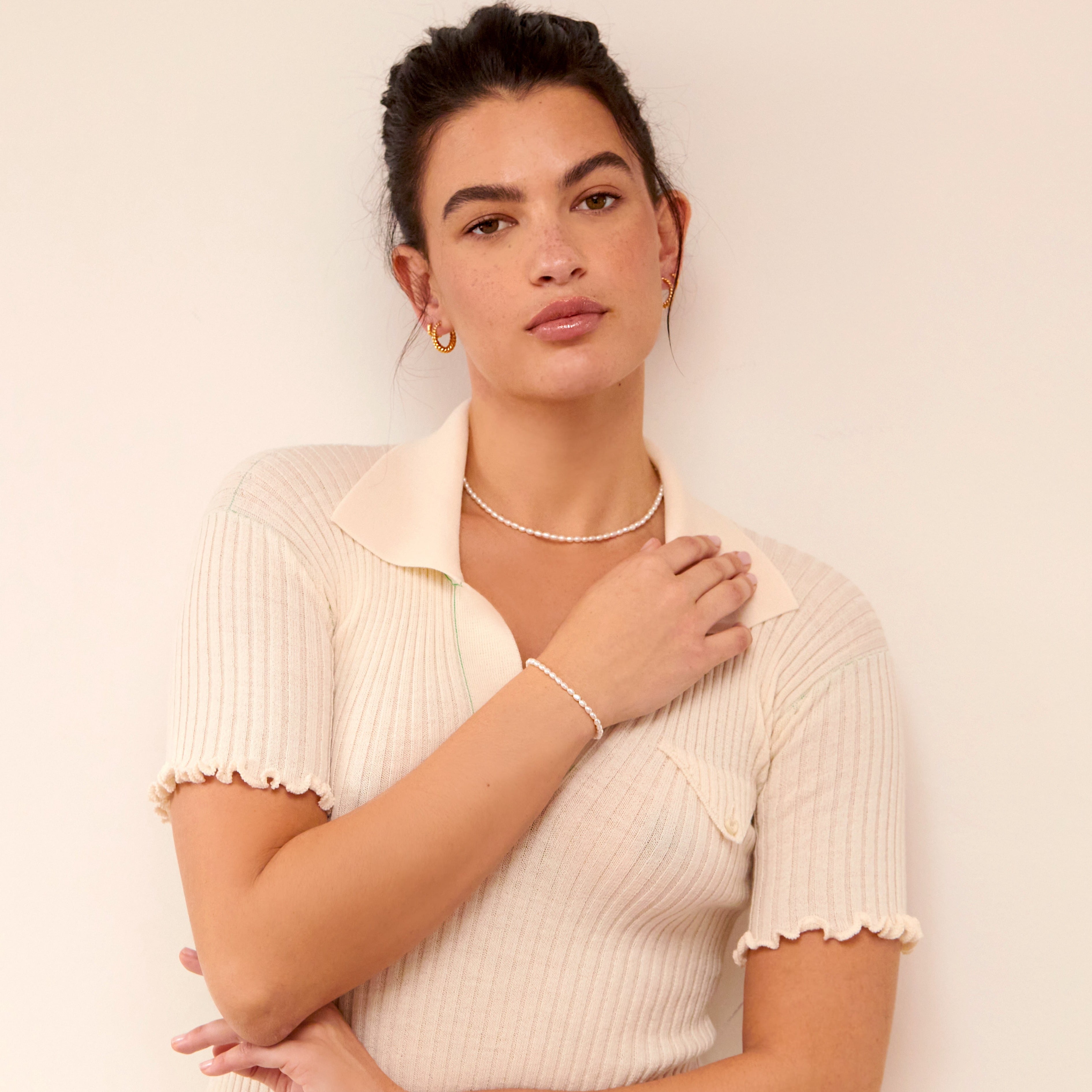 Woman wearing a silver seed pearl bracelet around her wrist, a silver seed pearl chocker around her neck, gold large twisted rope hoop earrings in her ear lobes and a cream knitted collared T-shirt