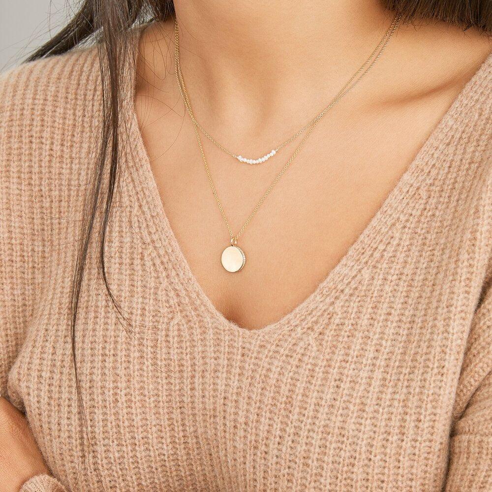 Plain gold small diamond style disc necklace layered with a gold small pearl cluster choker around the neck of a woman wearing a brown knitted sweater