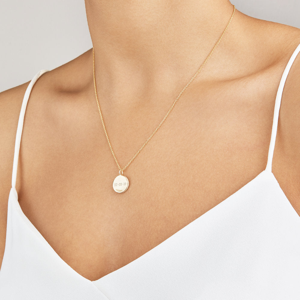 Gold small diamond style disc necklace with a date engraved on it around the neck of a woman wearing a white strappy top