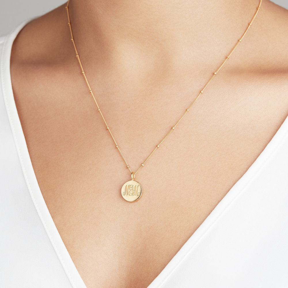 Gold satellite chain necklace with a circular gold pendant that has the letters 'N&M' engraved around the neck of a woman wearing a white v neck top