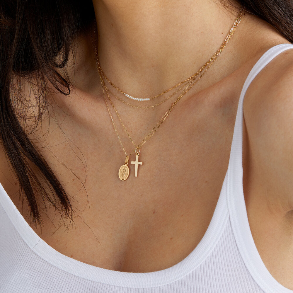 A gold cross necklace layered with a gold pearl cluster necklace and gold symbol necklace worn around the neck of a woman wearing a white vest-top