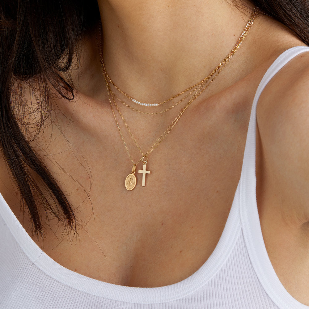 Solid gold cross necklace layered with a gold St  Christopher medallion necklace and a gold pearl cluster necklace around the neck of a woman wearing a white top