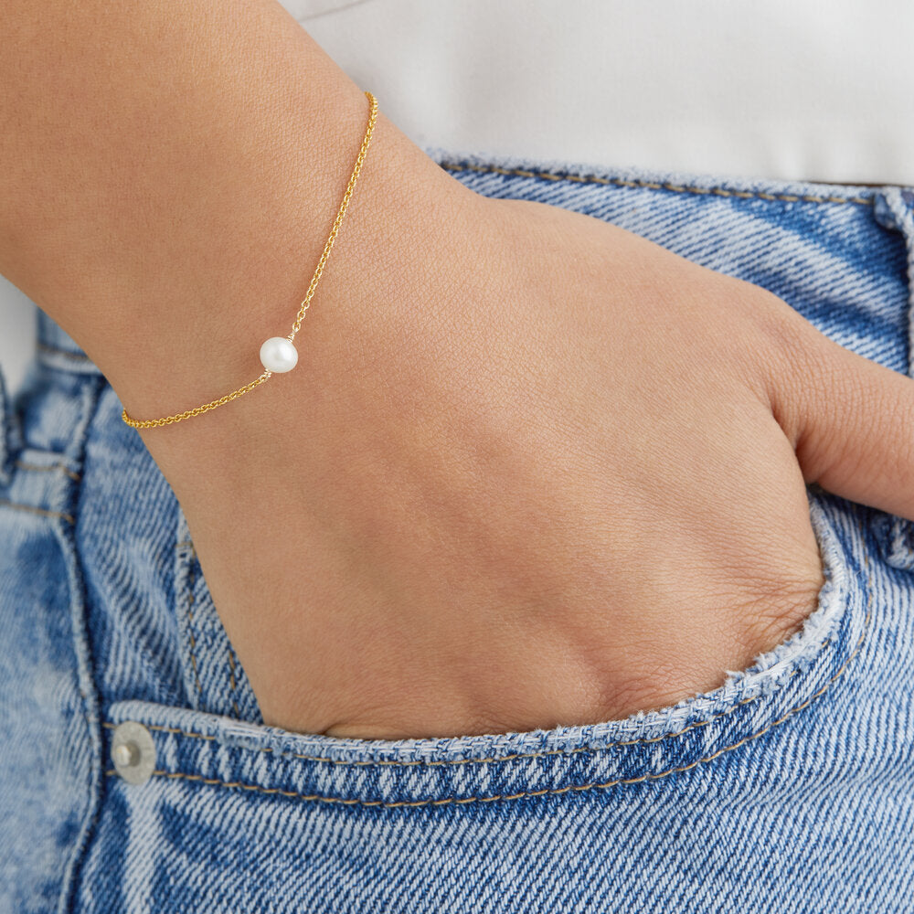 Gold single pearl bracelet around a wrist with the fingers in a denim blue pocket