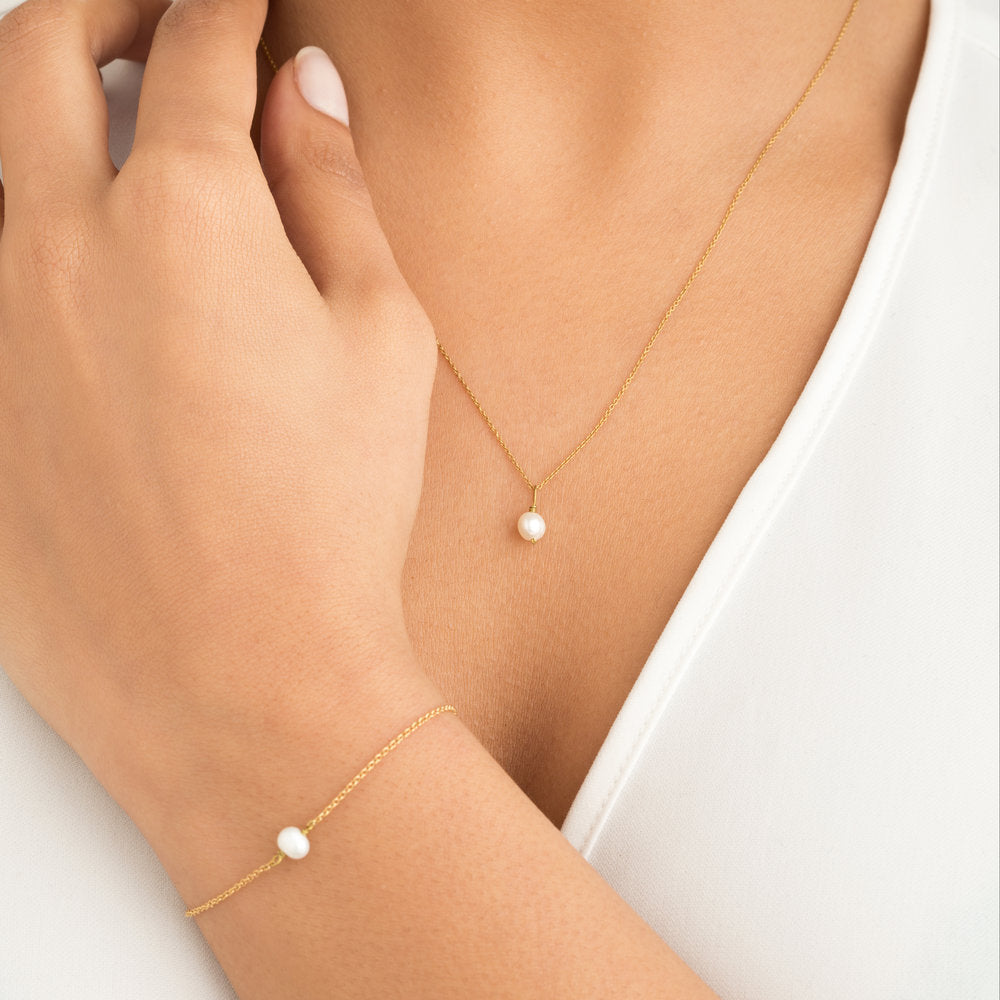 Gold single pearl necklace around the neck of a woman also wearing a gold single pearl bracelet and a white top