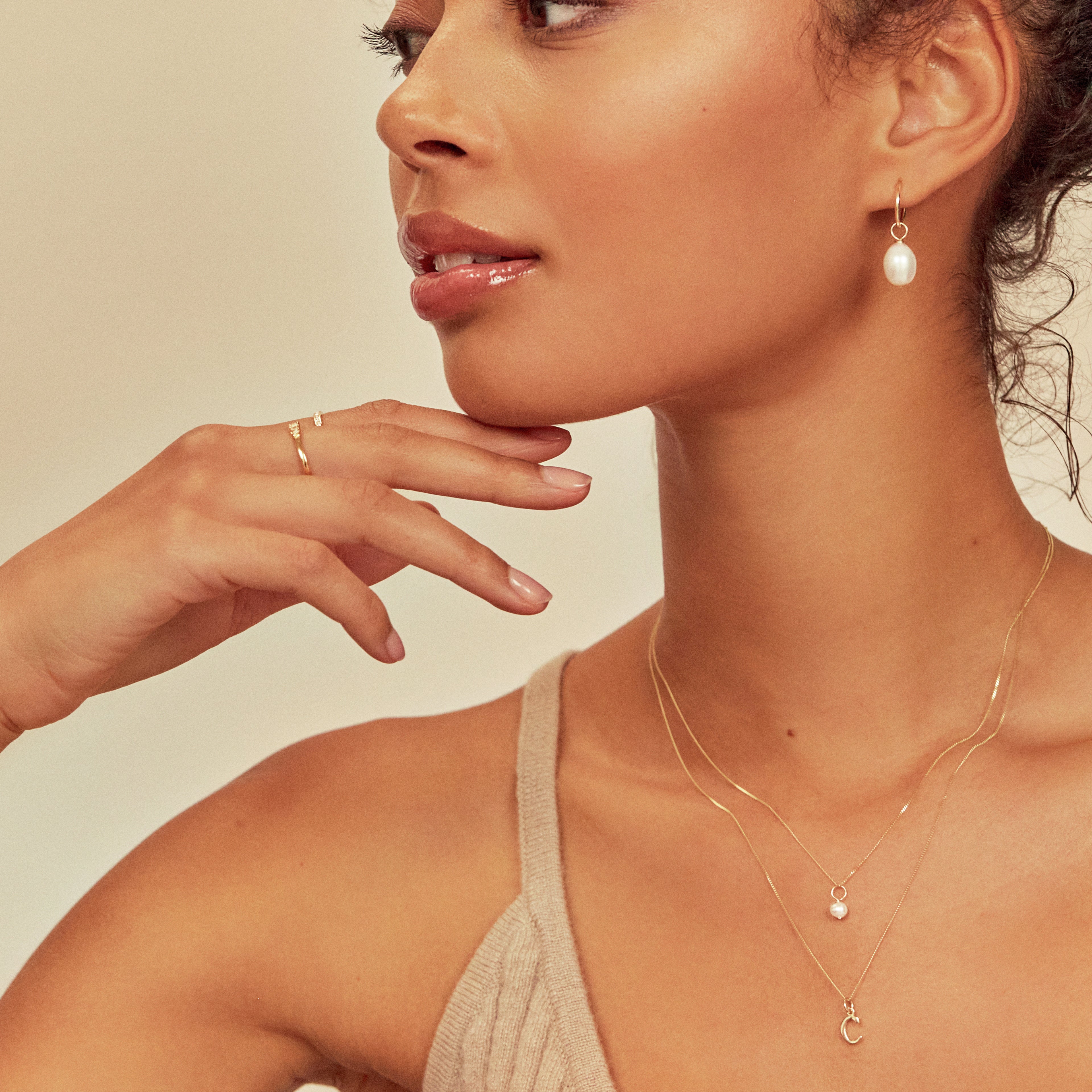 Gold single pearl necklace layered with a gold initial charm necklace 'C' around the neck of a woman  also wearing gold large pearl drop earrings in her ears and a brown knitted strappy top