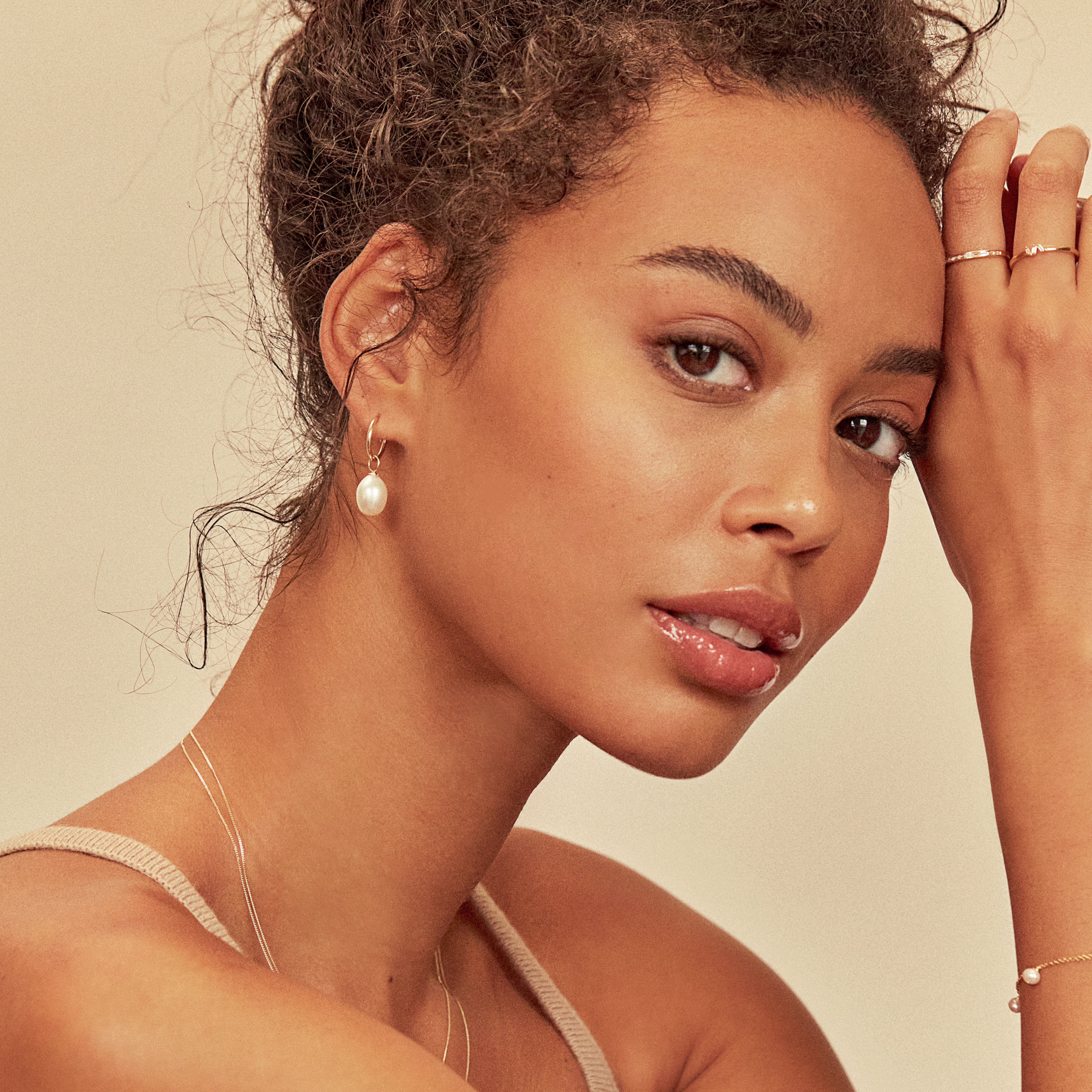 Solid gold large pearl drop hoop earring in an earlobe of a curly haired woman with her forehead resting on her hands