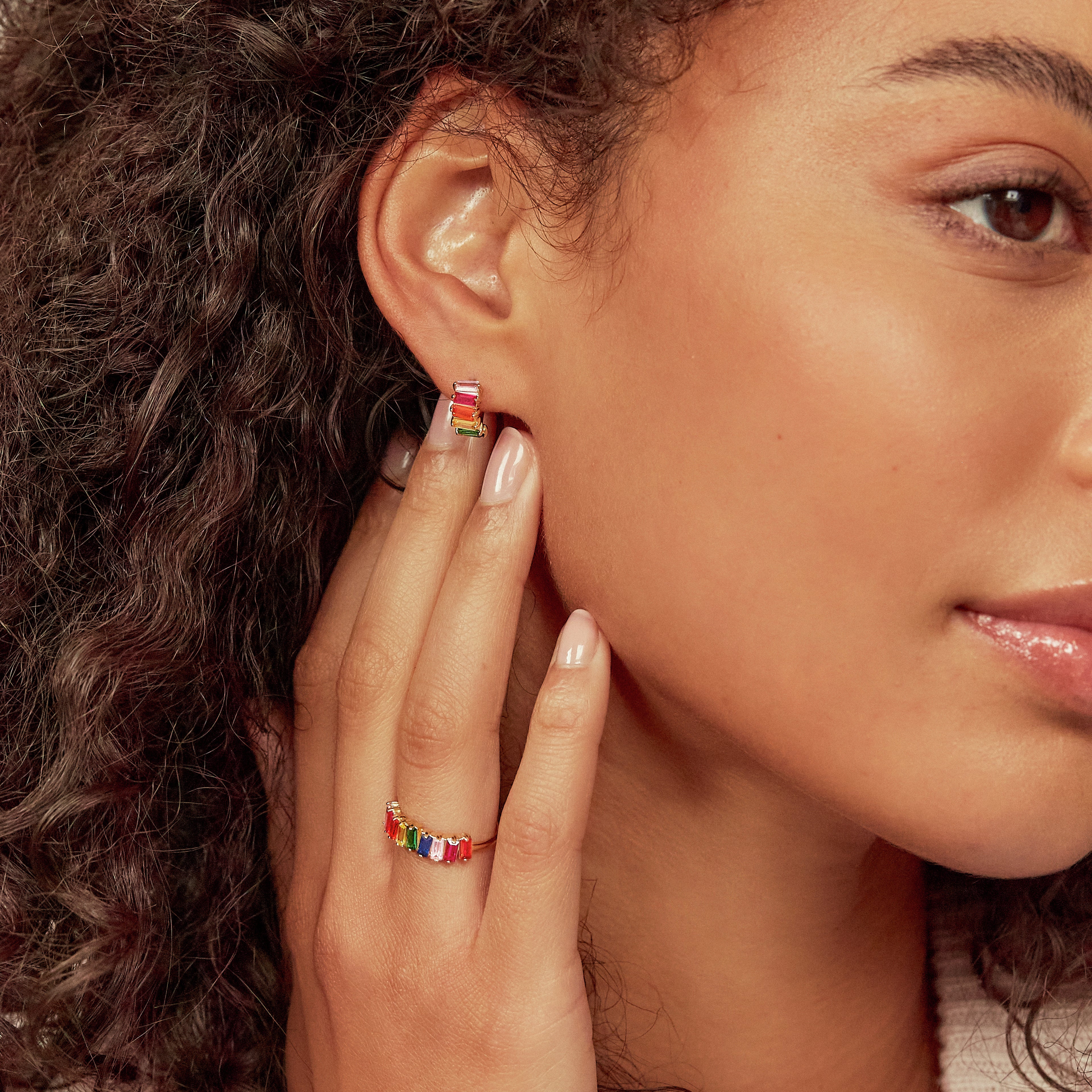 Gold rainbow gemstone cluster huggie hoop earring in the ear lobe of a woman wearing a gold rainbow gemstone cluster ring on her finger