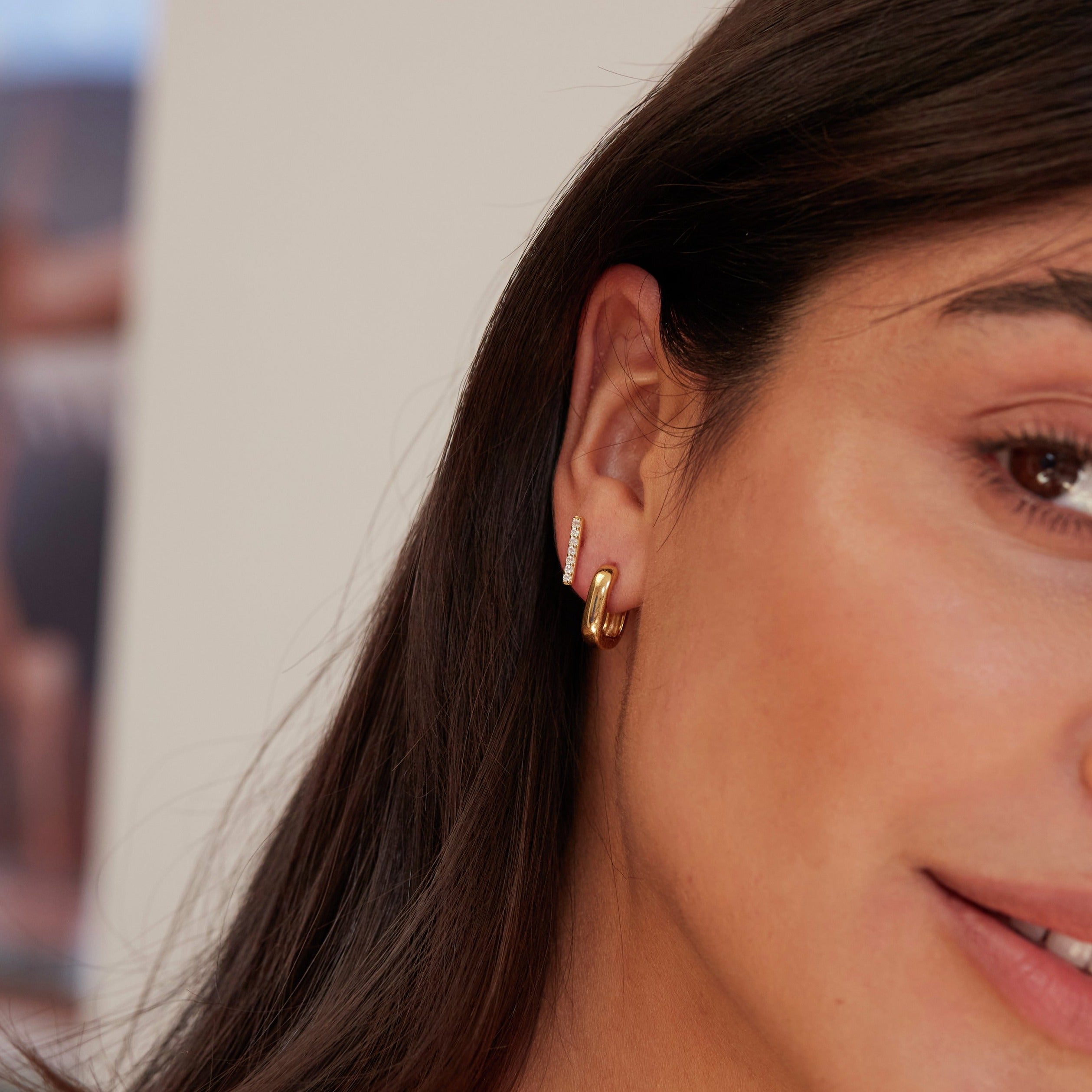 A brunette woman wearing a gold thick squared hoop earring and a gold diamond style bar stud in her ear lobe