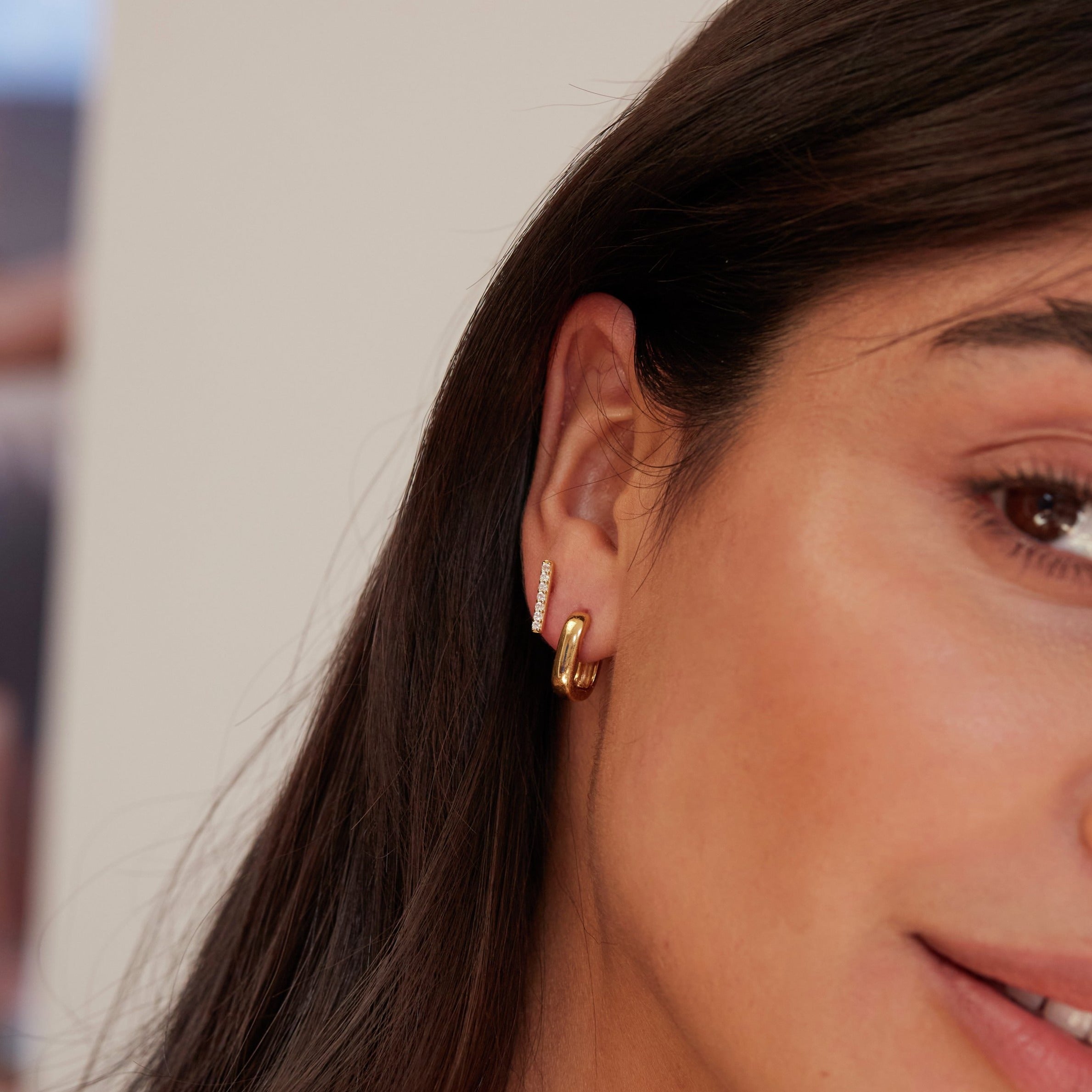 A gold thick squared hoop earring and a gold diamond style bar stud in an ear lobe