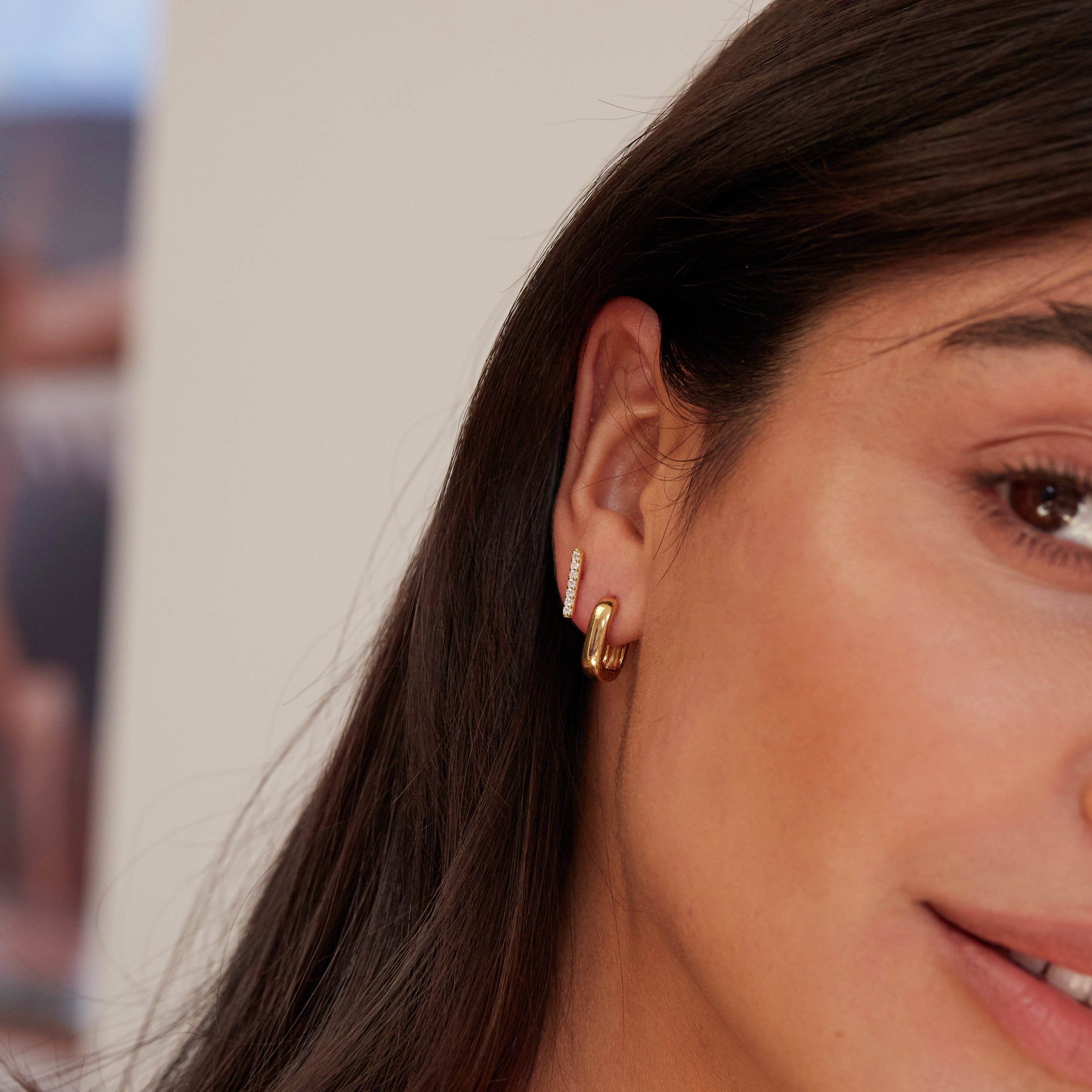 Gold thick squared hoop earring paired with a diamond earring in one ear lobe of a brunette woman 