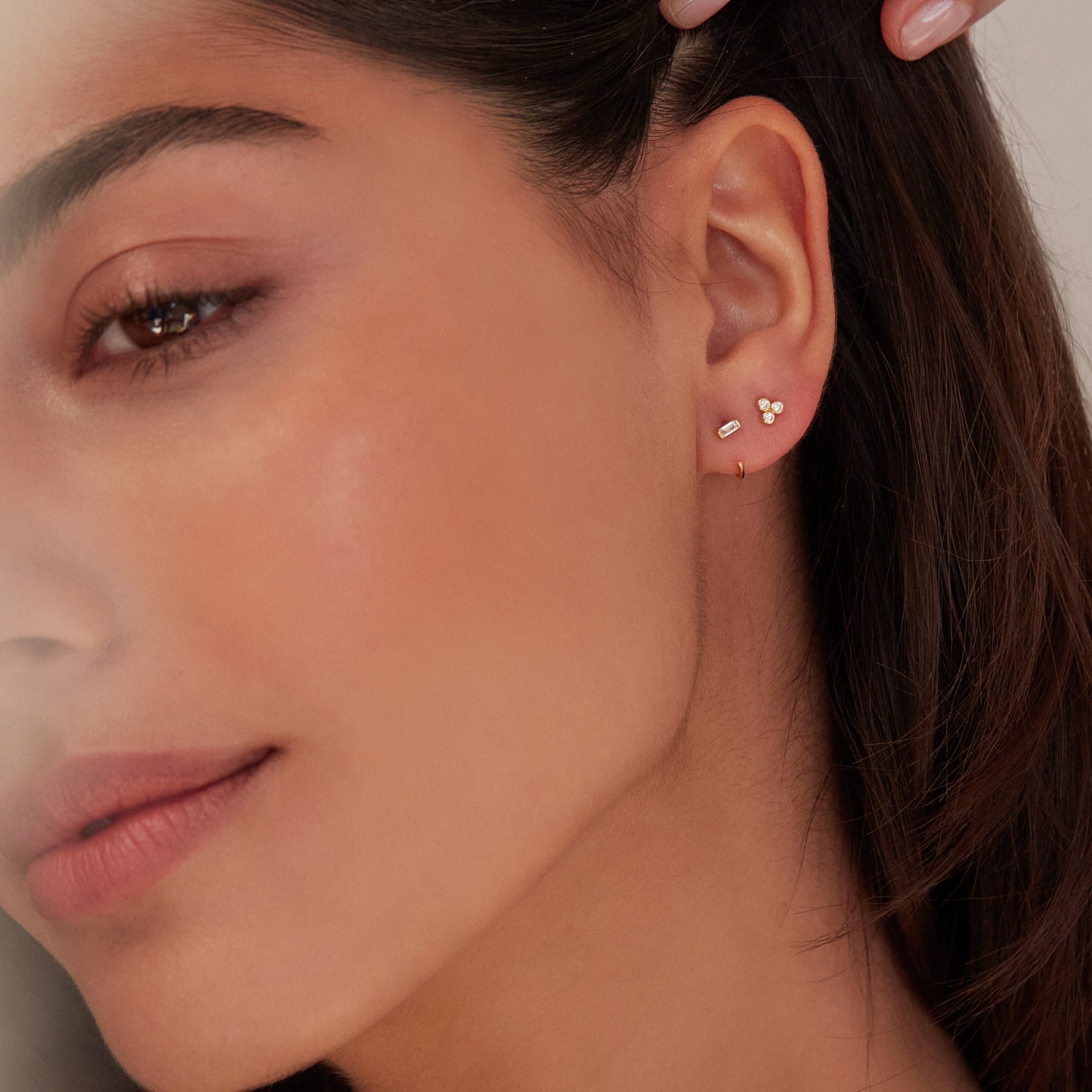 A gold diamond style pyramid stud earring and a gold diamond style lobe hoop stud earring in an ear lobe of a brunette woman