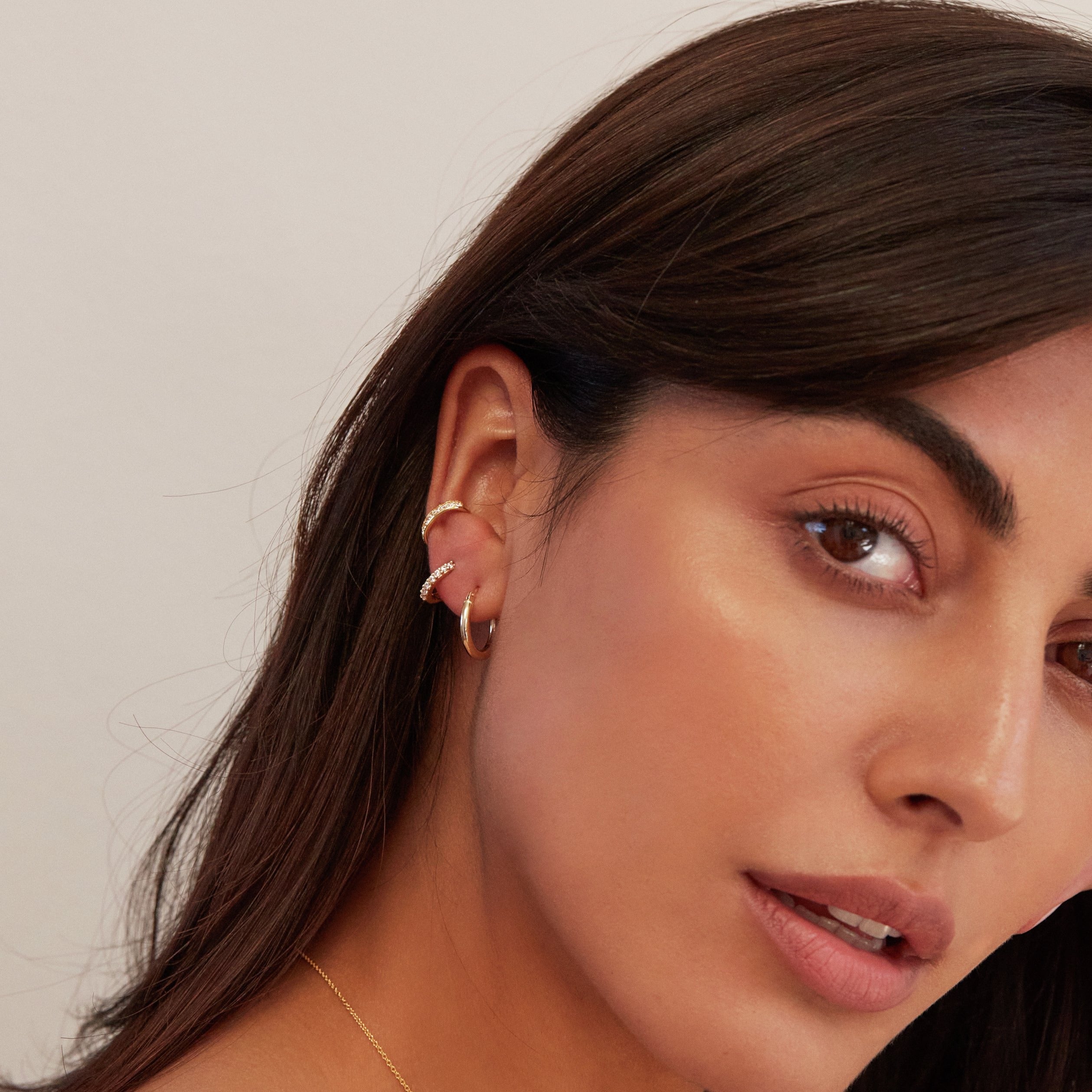 Gold thick diamond style ear cuff in one ear of a brunette woman wearing another gold cuff and a gold huggie hoop earring in her ear