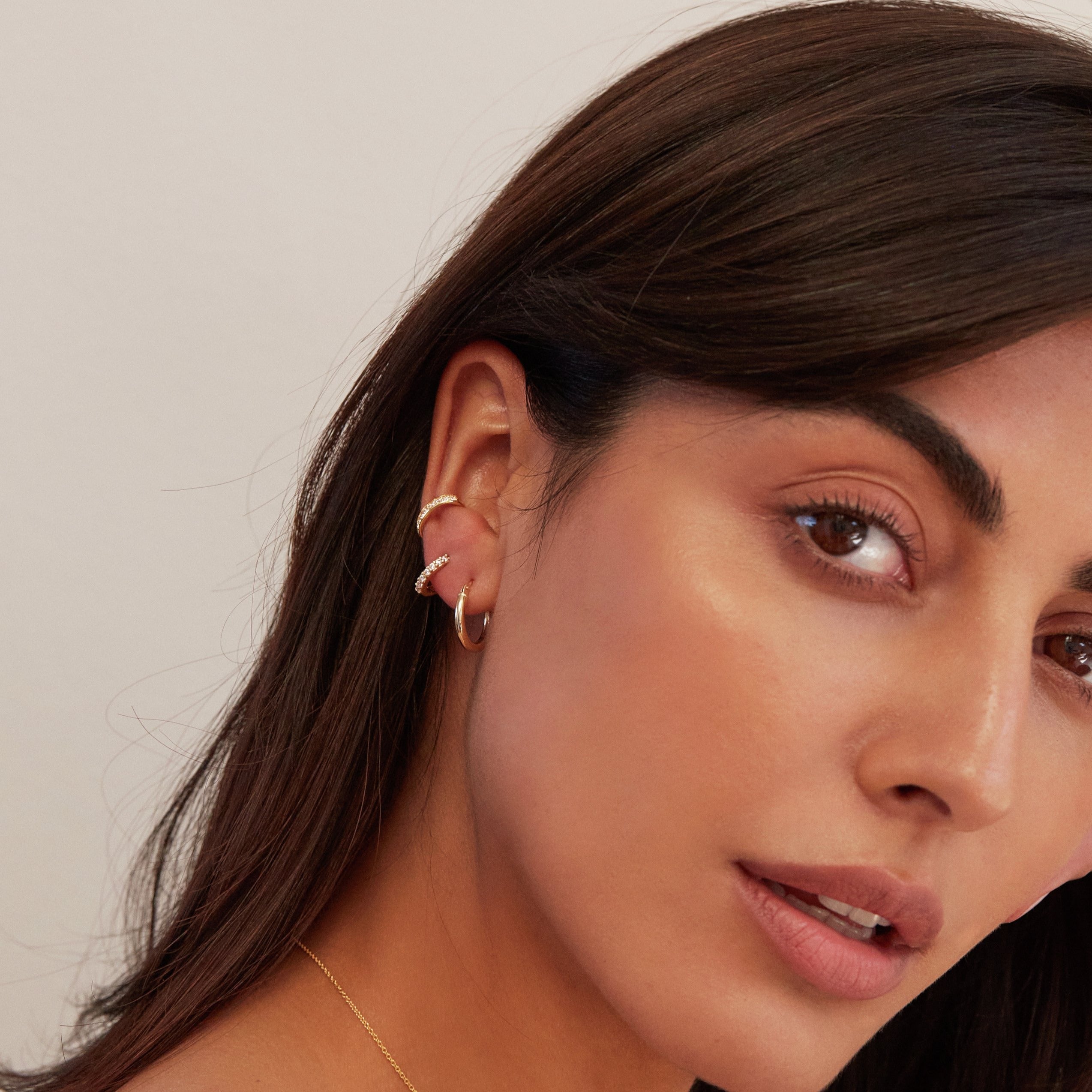 A brunette woman wearing two gold thick diamond style ear cuffs and a gold huggie hoop earring in her ear lobe