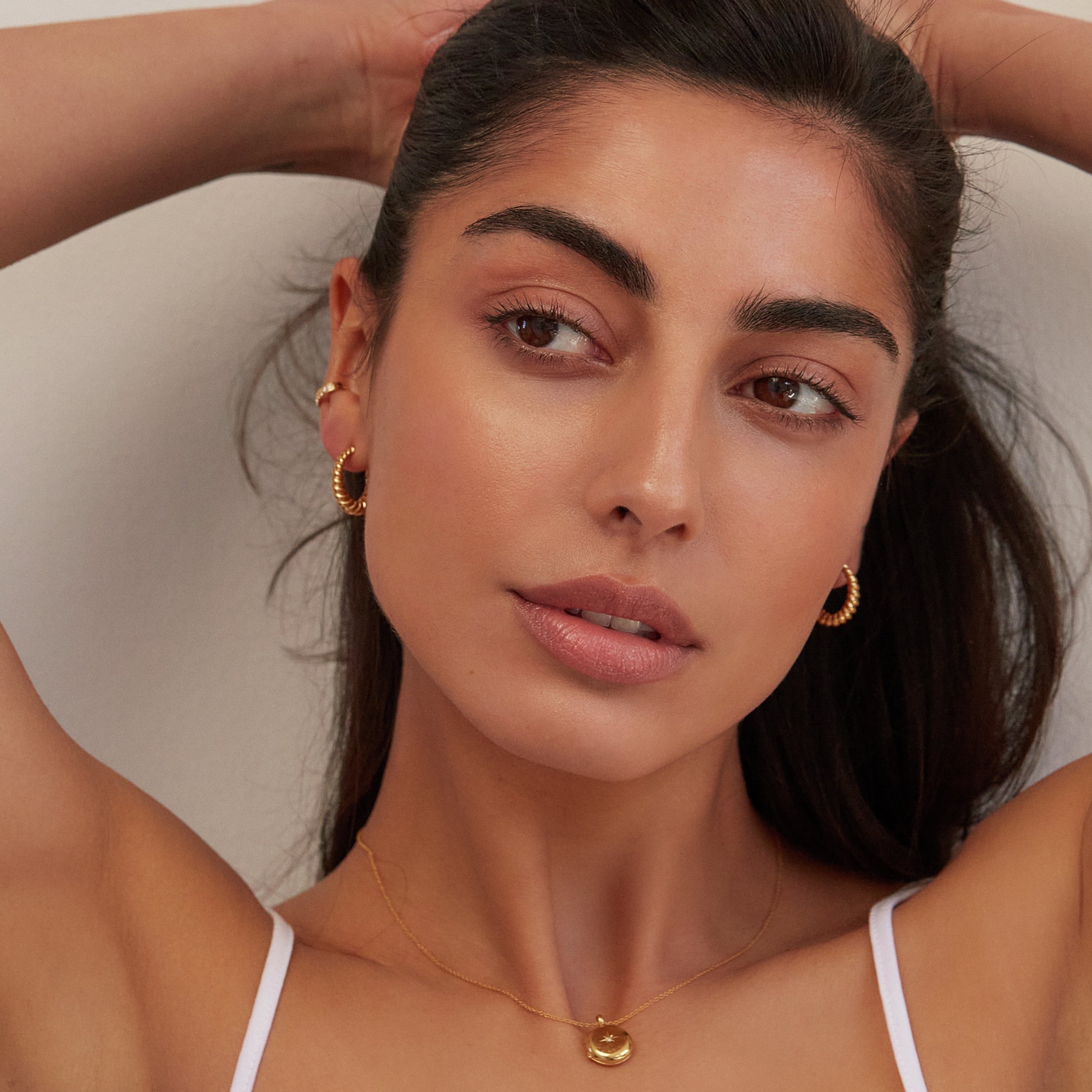 Gold large twisted rope hoop earrings worn by a brunette woman holding her hair tightly back with her hands also wearing a gold locket necklace
