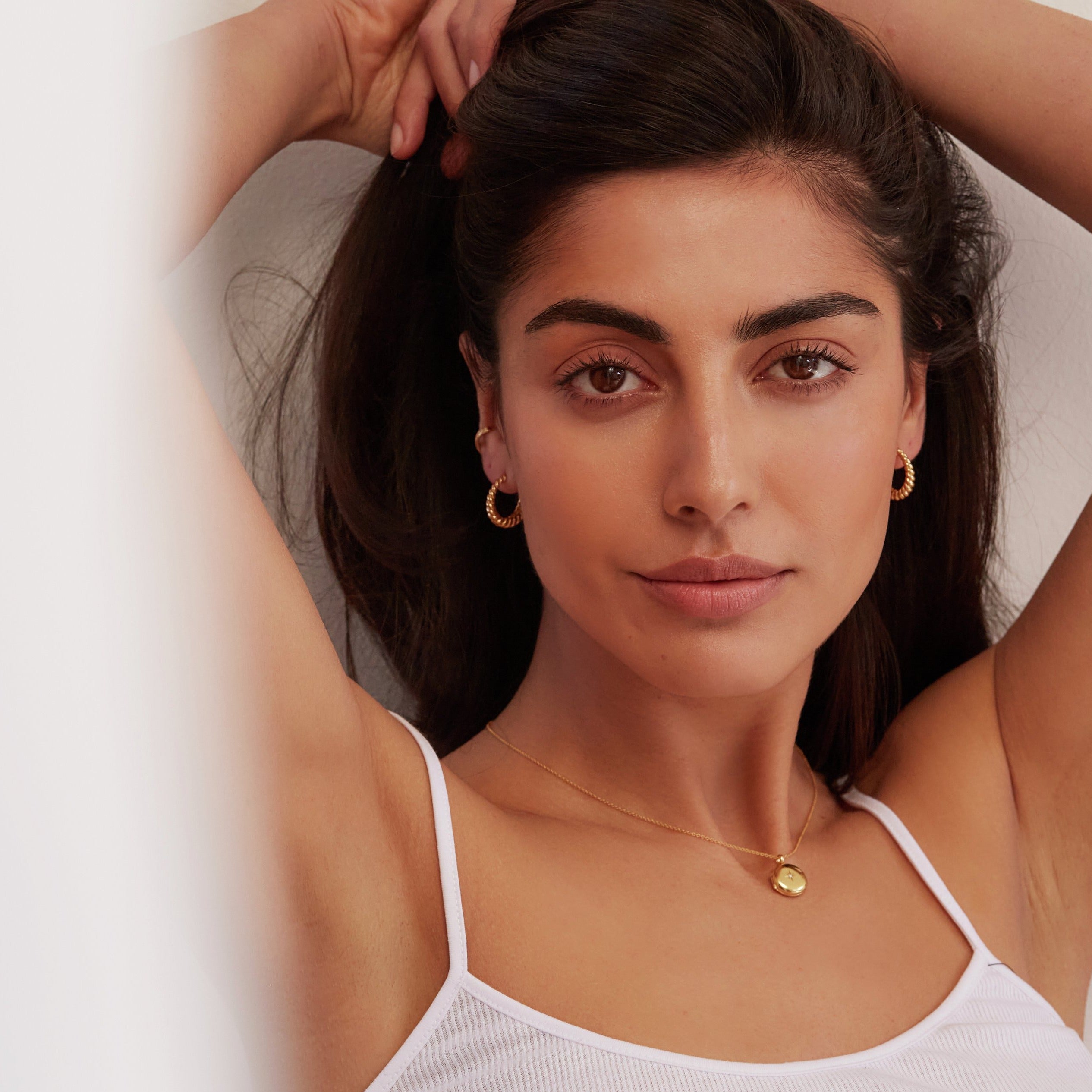 Gold large twisted rope hoop earrings worn by a brunette woman holding her hair back with her hands also wearing a gold small round diamond locket necklace