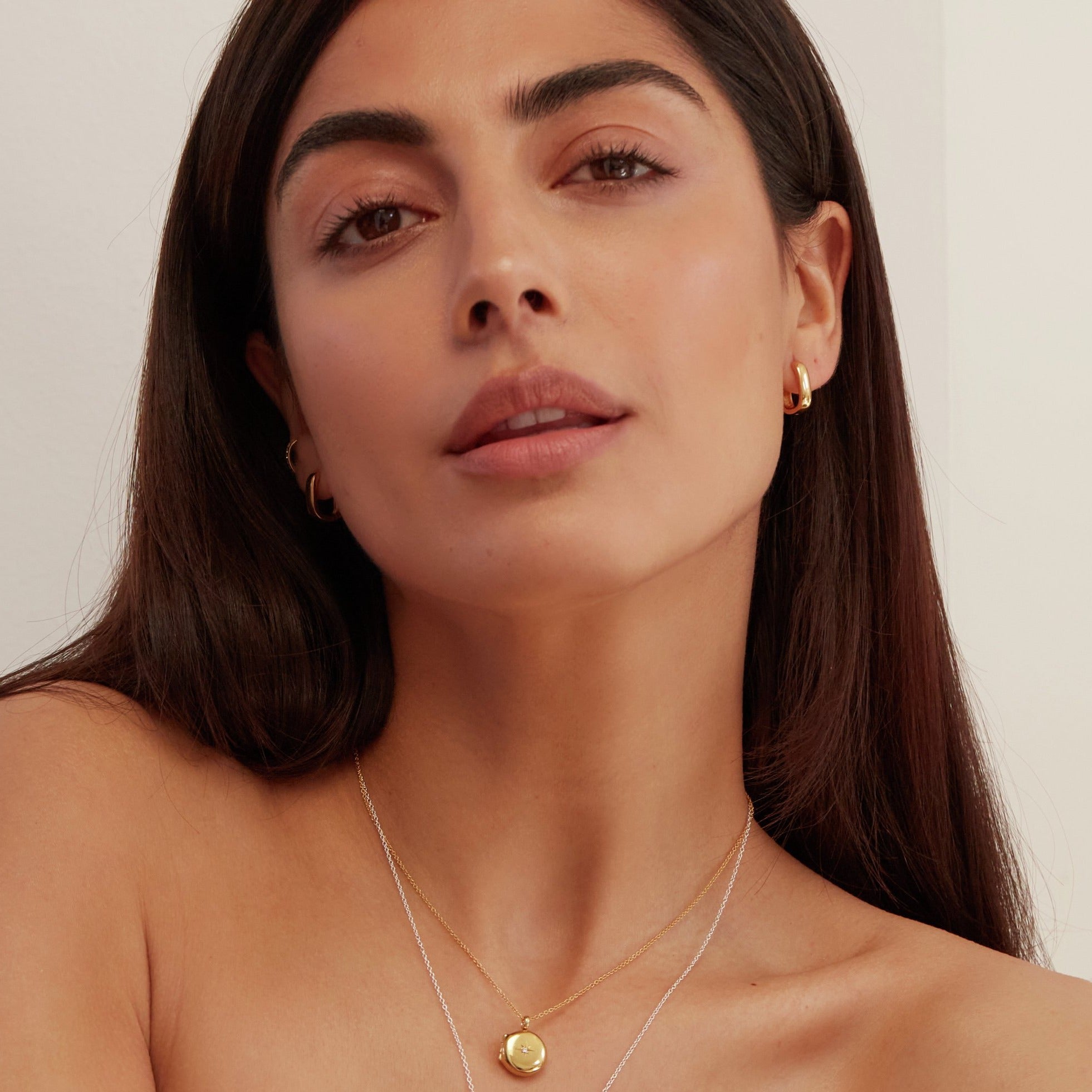 Gold thick squared hoop earrings in the ear lobes of a brunette woman wearing a gold small round diamond locket necklace around her neck