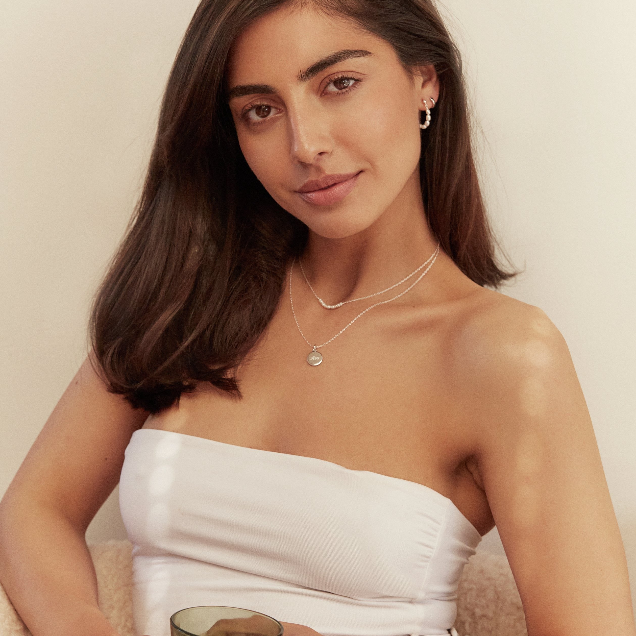 A woman wearing a silver small round engraved disc necklace with the name 'Ava' engraved and a silver pearl cluster choker around her neck and a white strapless top on her body