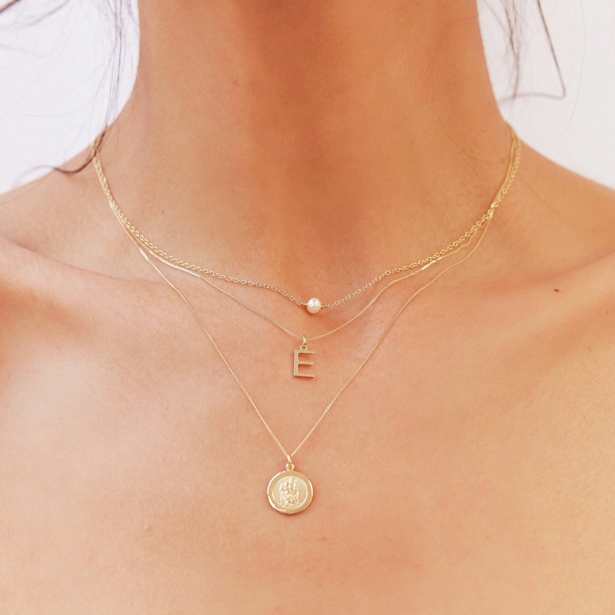Close up upward angle shot of a neck wearing a gold single pearl choker layered with a gold E letter necklace and gold symbol necklace