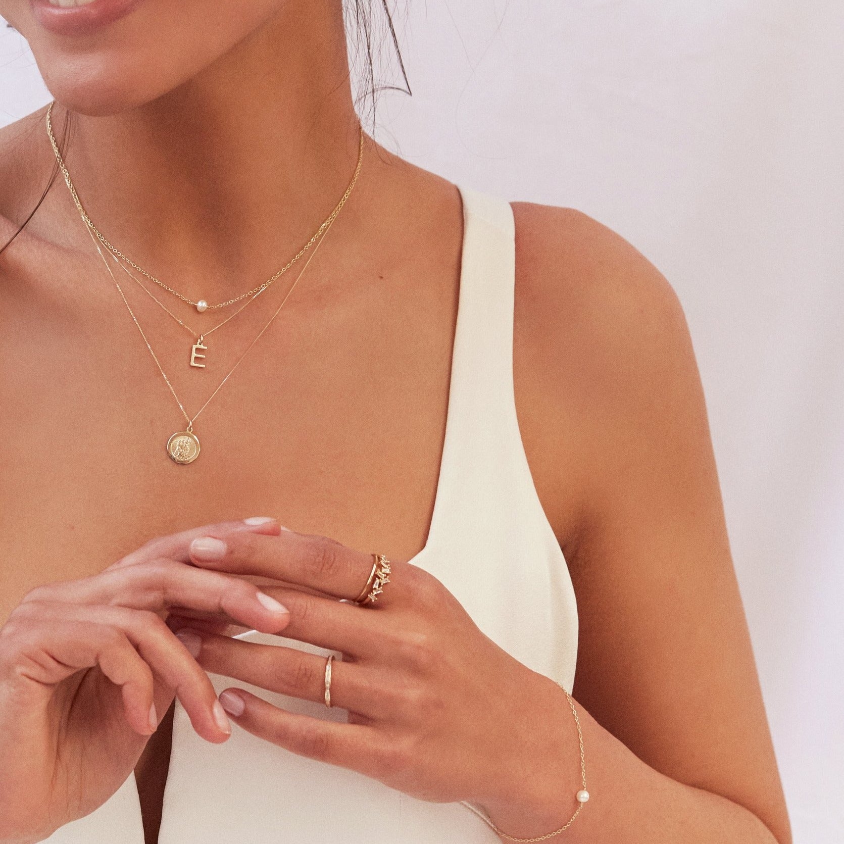 A woman wearing a gold single pearl bracelet on a wrist her wrist with a gold diamond style baguette ring on her finger, a gold single pearl choker around her neck layered with a gold E letter necklace and a gold symbol necklace
