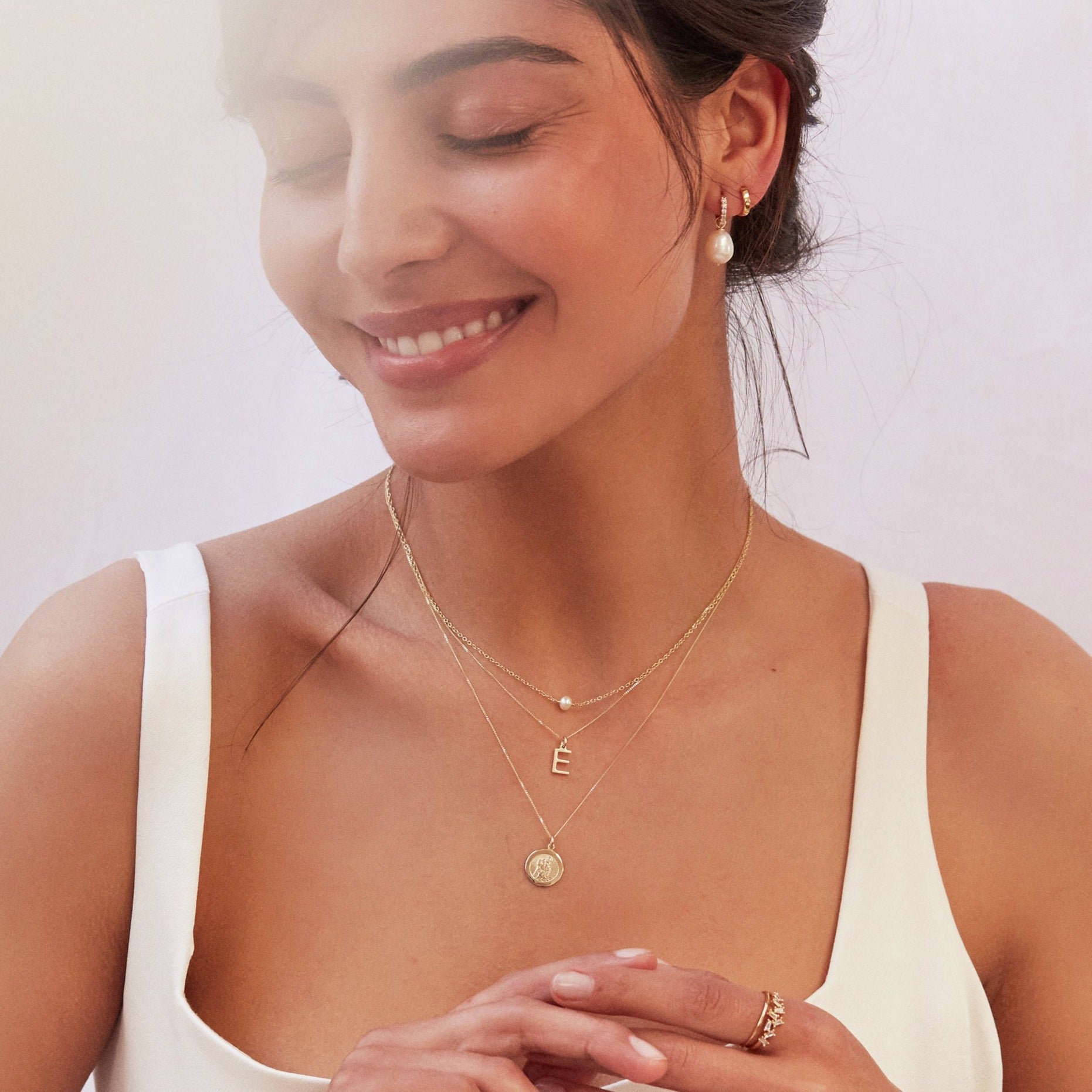 Smiling brunette woman wearing a gold single pearl choker around her neck layered with a gold E letter necklace and gold symbol necklace 