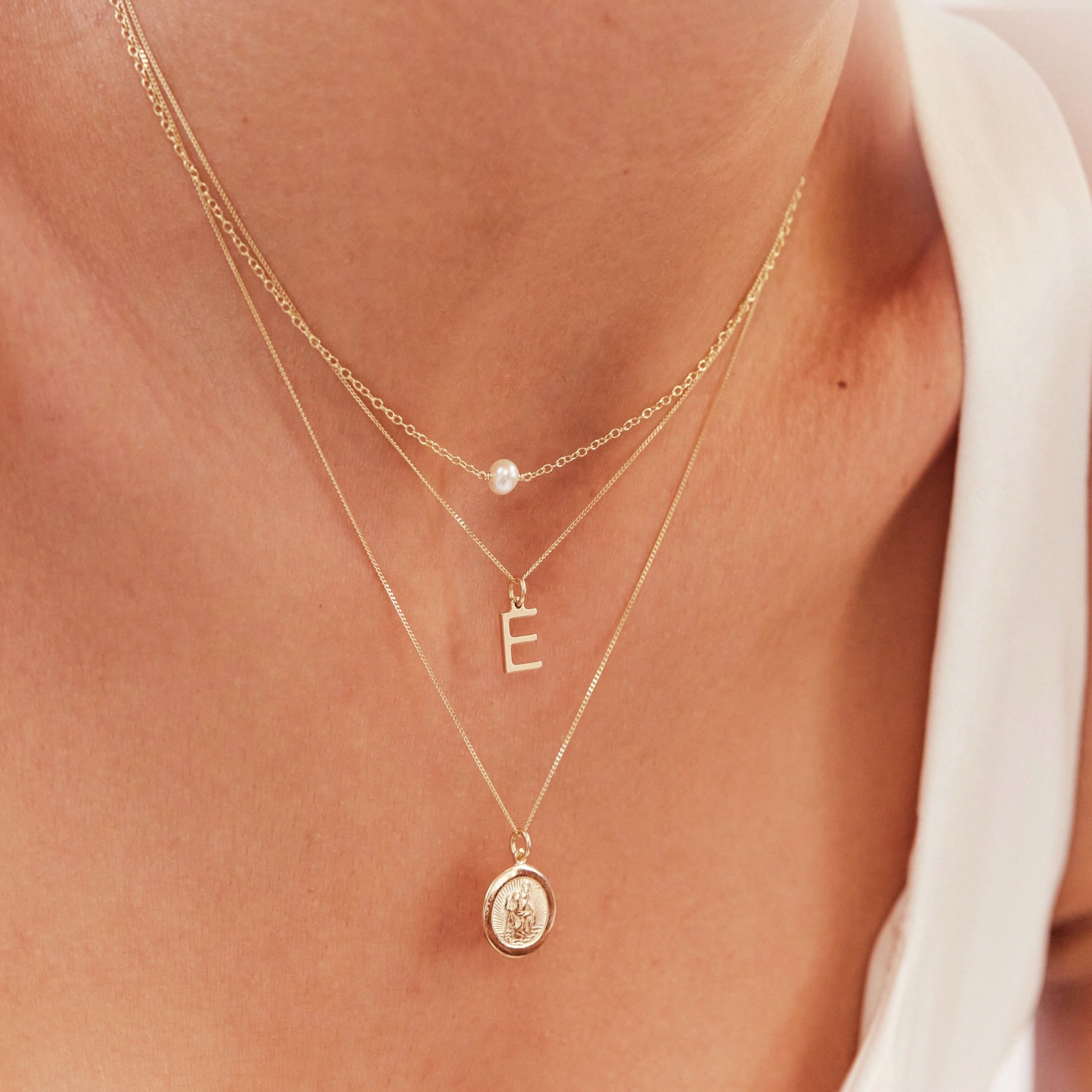 Close up of a neck wearing a gold single pearl choker layered with a gold E letter necklace and gold symbol necklace