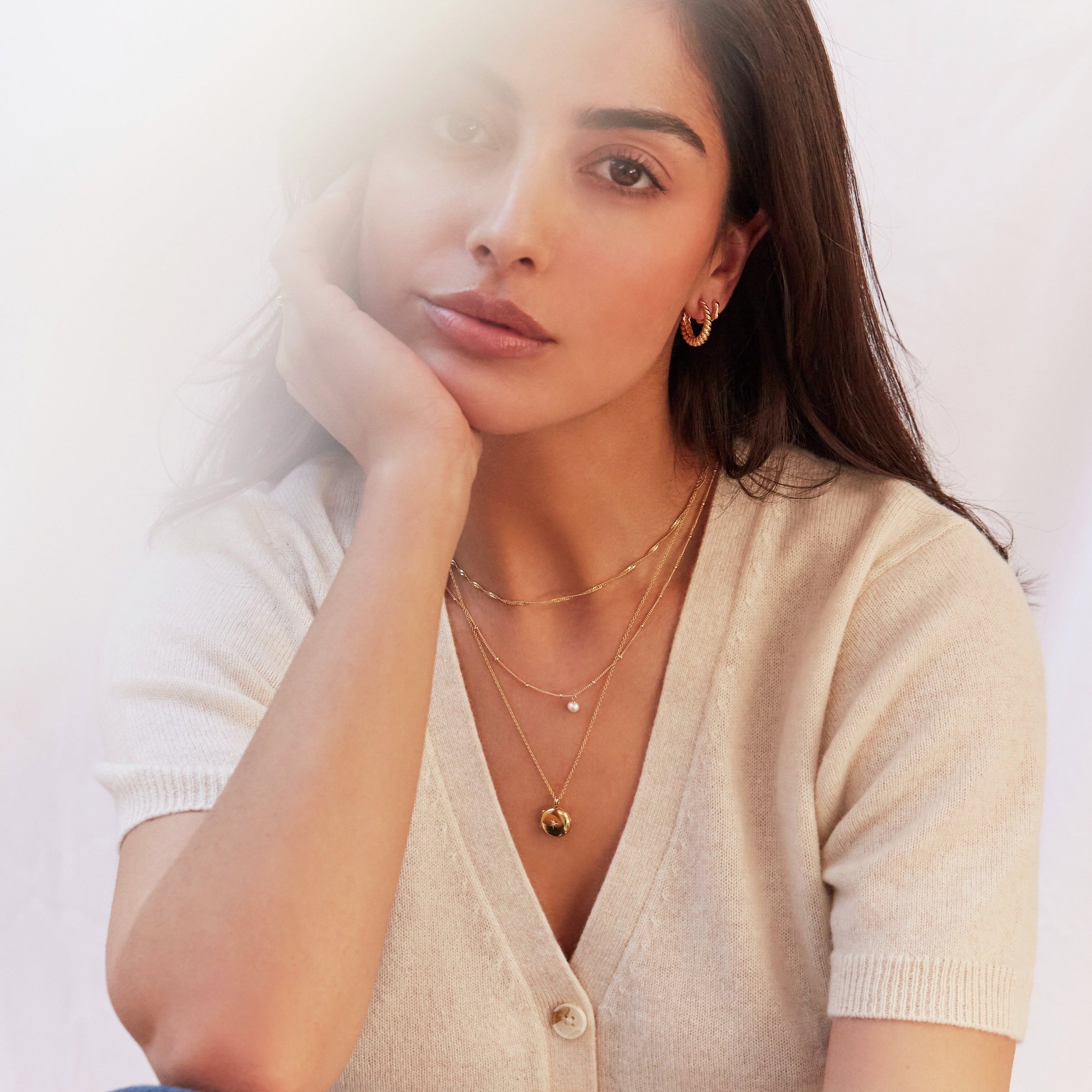 Gold large twisted rope hoop earring and gold molten huggie hoop earring in one ear lobe of a brunette woman with her hand holding her face wearing a gold small round diamond locket necklace and more gold necklaces