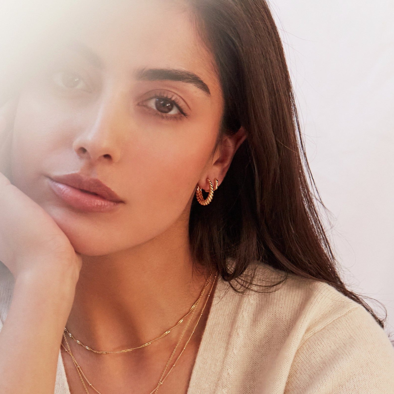 Gold large twisted rope hoop earring and gold molten huggie hoop earring in one ear lobe of a brunette woman with her hand holding her face wearing a gold small round diamond locket necklace and more gold necklaces