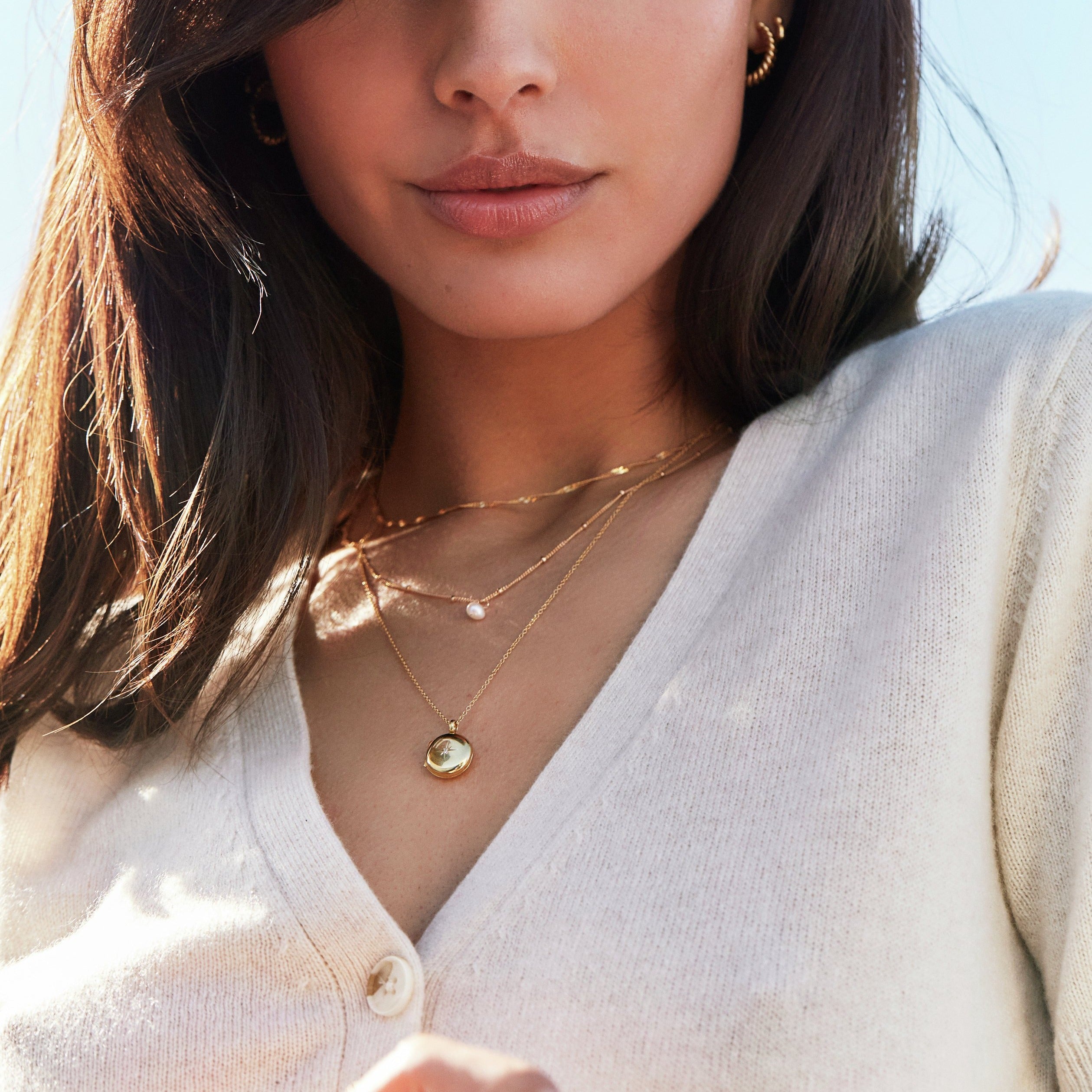 A woman wearing a gold small round diamond locket necklace, a gold twisted rope chain necklace and a gold single pearl satellite necklace around her neck with a beige sweater and a gold large twisted rope hoop earring in her ear lobe