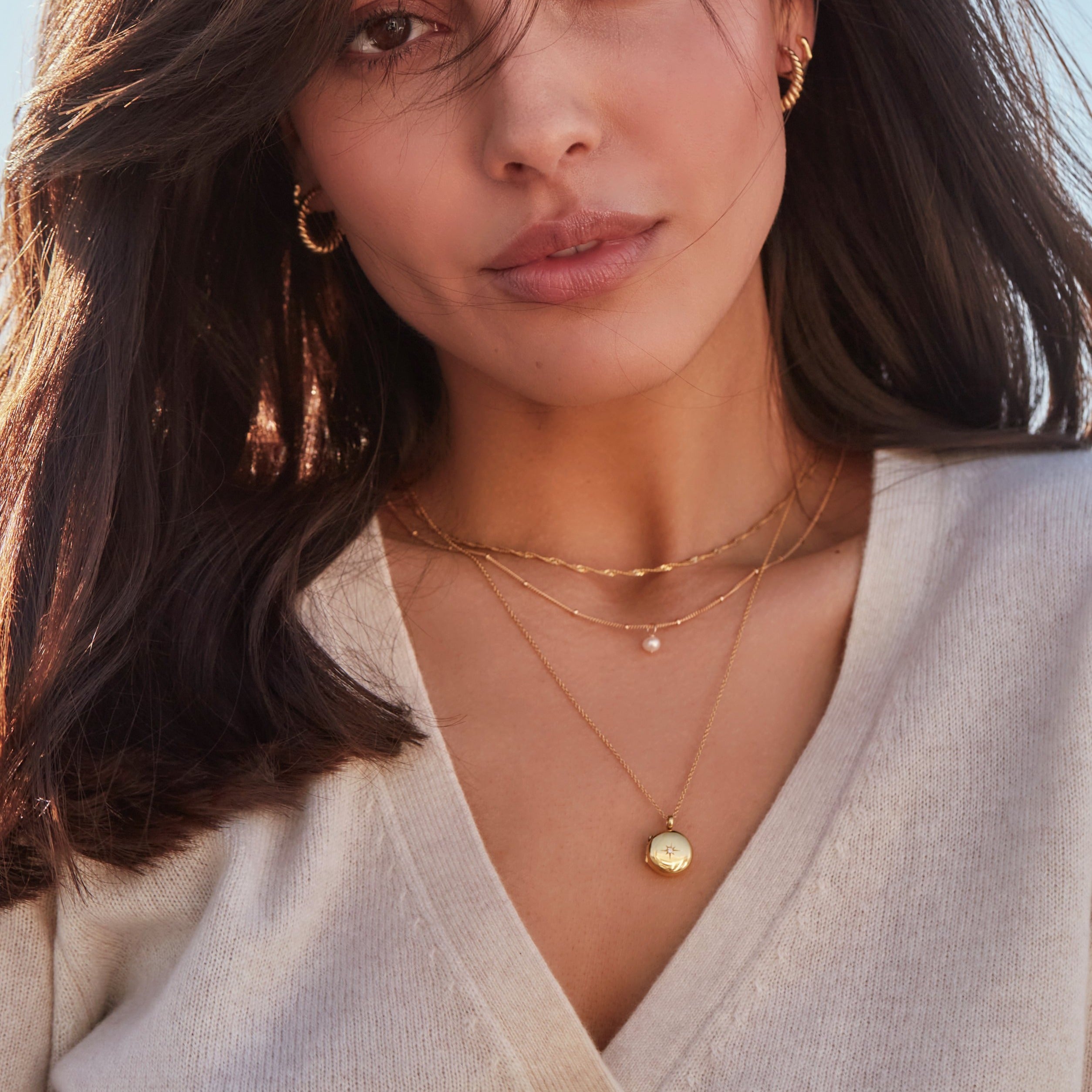 Brunette woman wearing a gold single pearl satellite necklace layered with a gold twisted rope necklace and gold small round diamond locket necklace with gold large twisted rope hoop earrings in her ears 